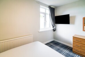 A room with a white bed, a radiator, a window with curtains, a wooden dresser, and a black television on the wall. - Adamton Country House Hotel (Prestwick)