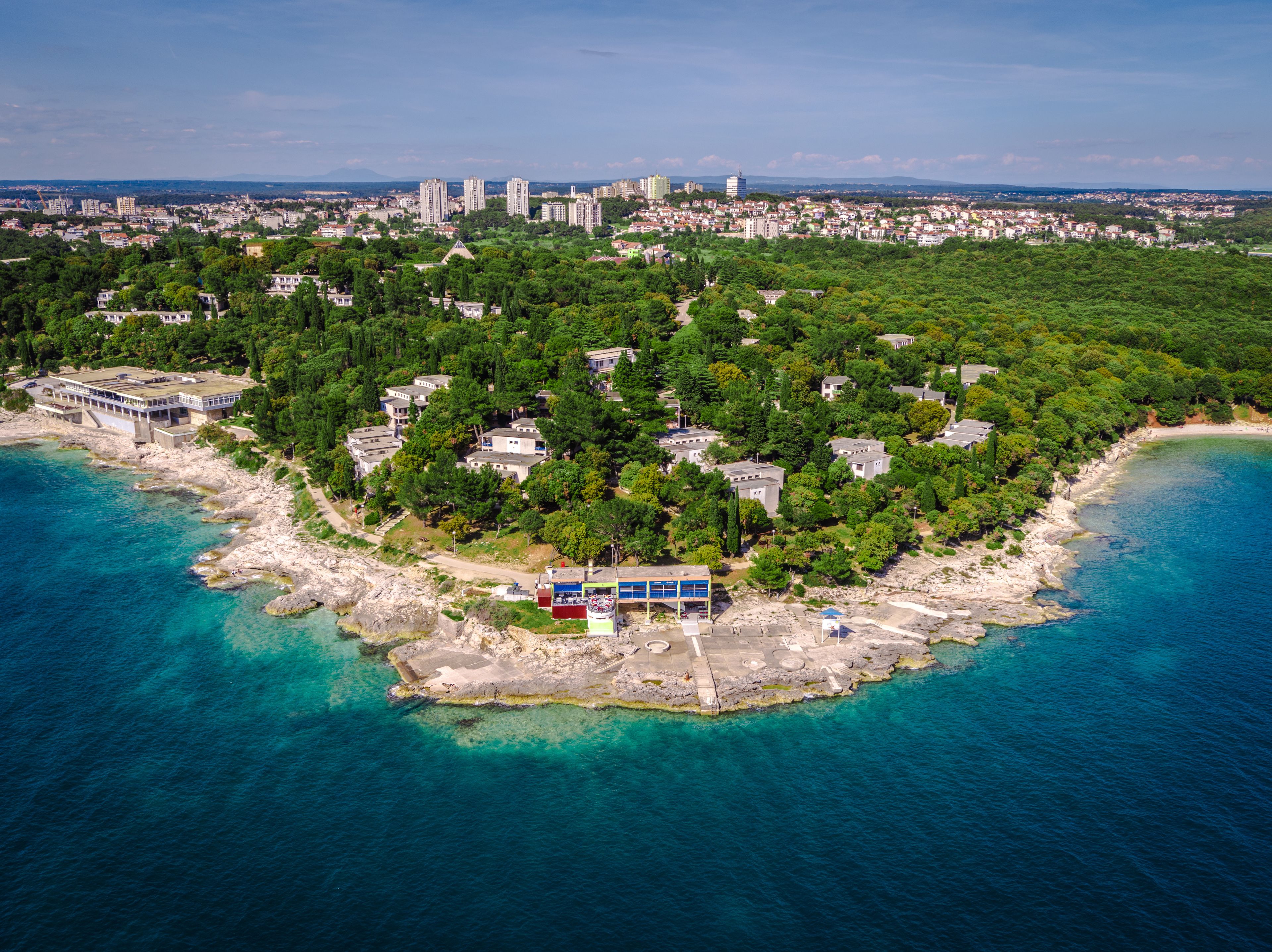 An aerial view of a coastal area with a mix of residential buildings and greenery, a rocky shoreline, and clear blue waters.