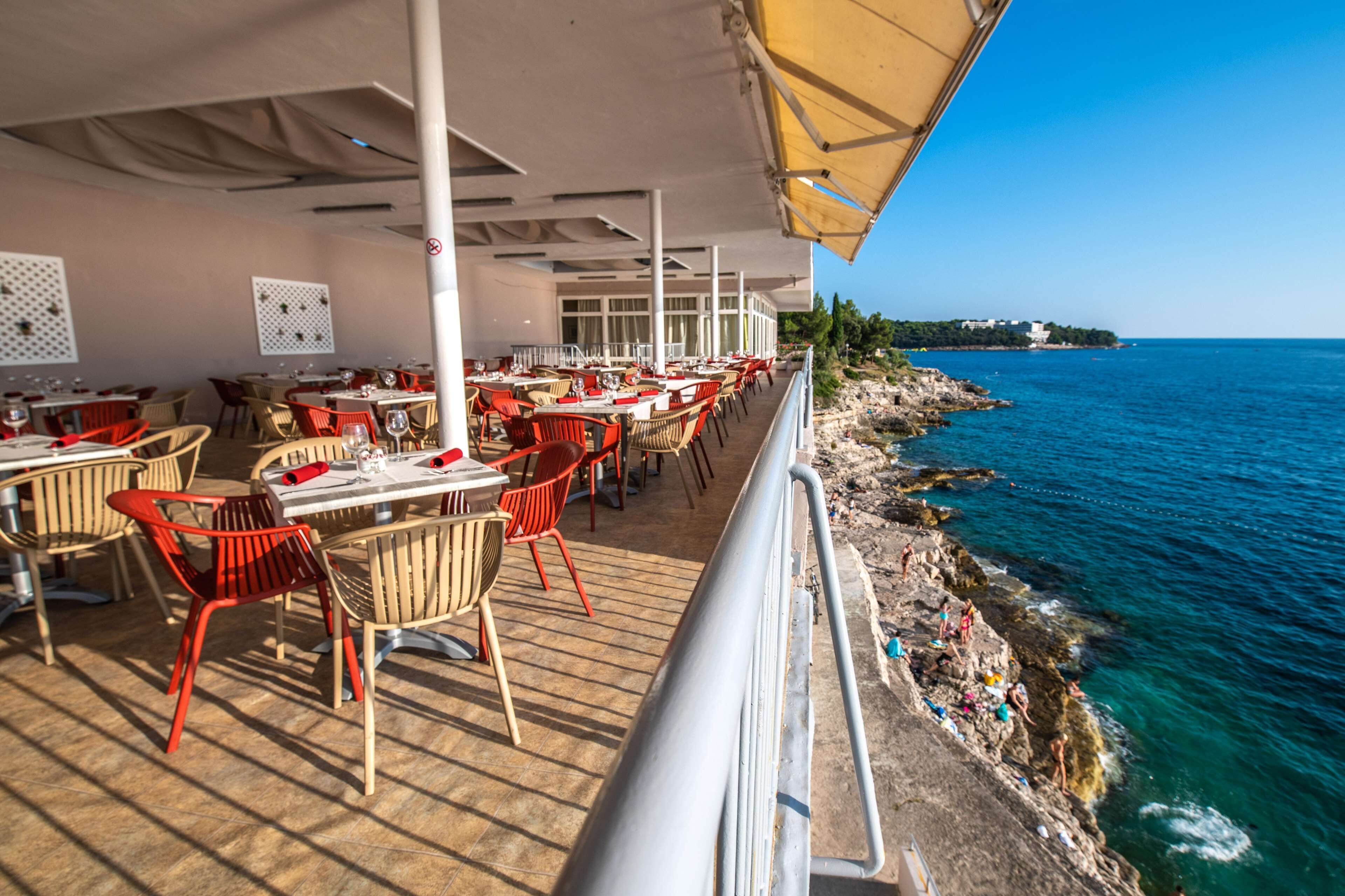 A seaside restaurant with outdoor seating, tables, and chairs, overlooking the sea.