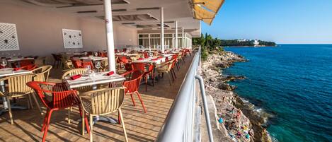 A seaside restaurant with outdoor seating, tables, and chairs, overlooking the sea.