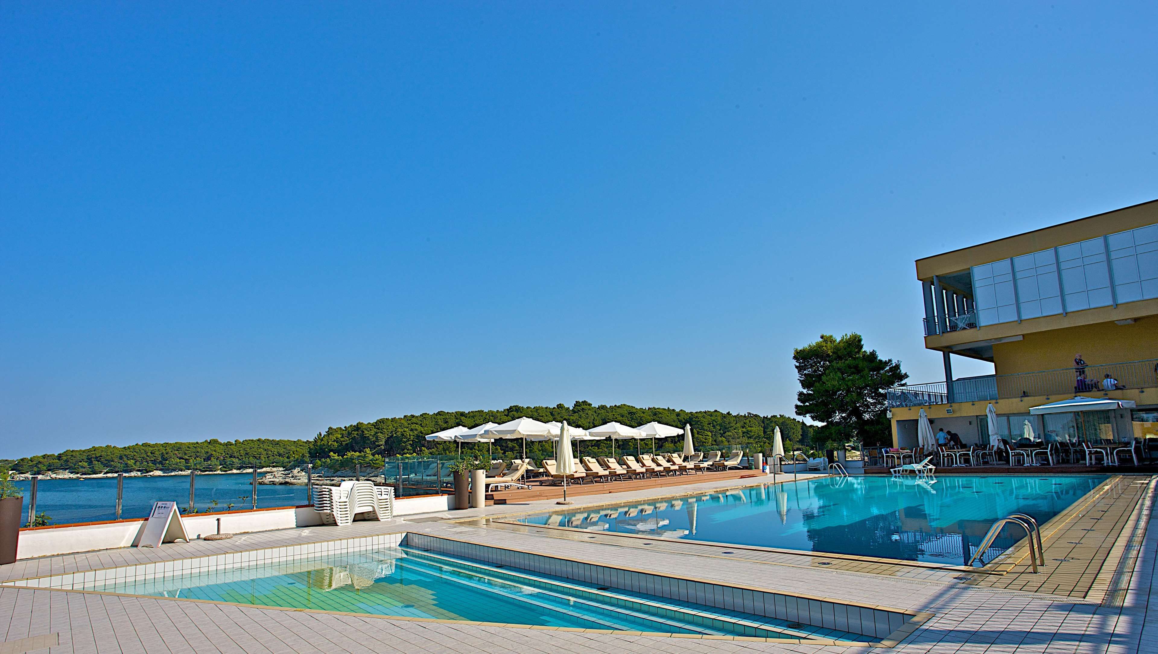 A pool area with lounge chairs and umbrellas, a clear view of the sea, and a building with a balcony.