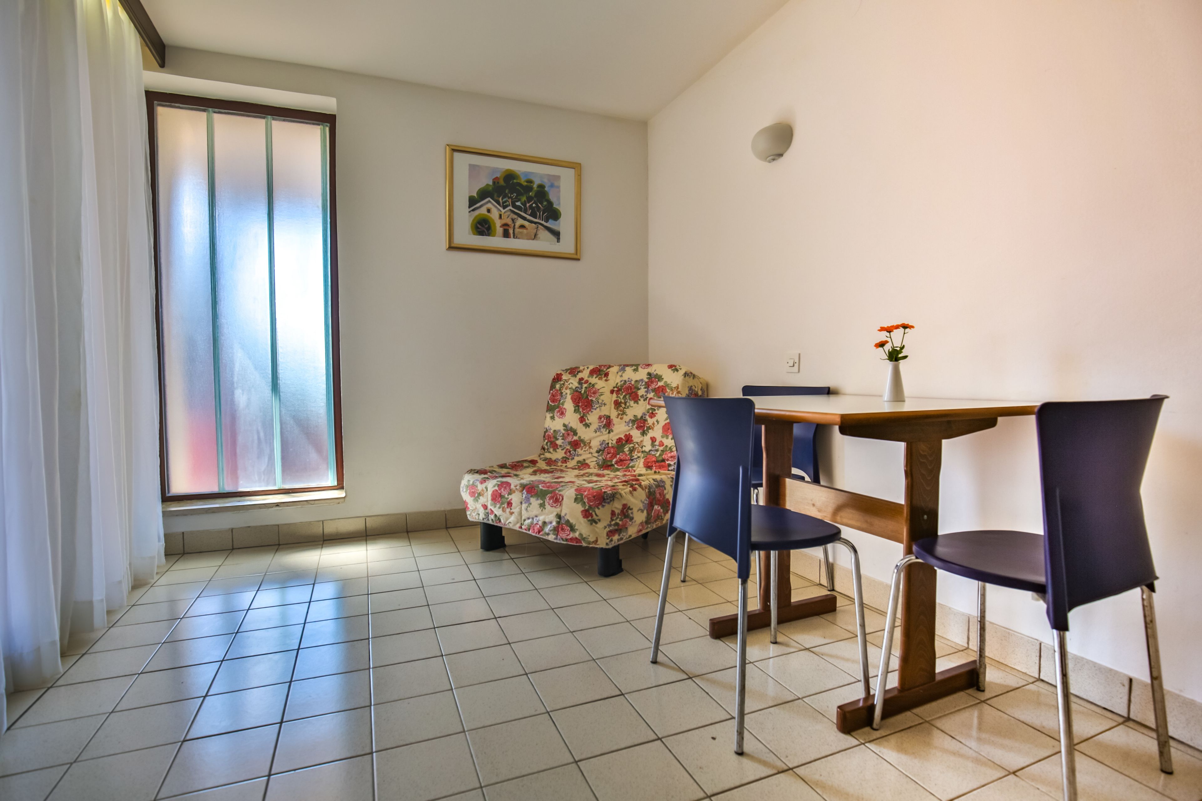 Un coin repas comprenant une chaise à motifs floraux, une table avec des chaises, une fenêtre avec des rideaux et un tableau encadré accroché au mur.