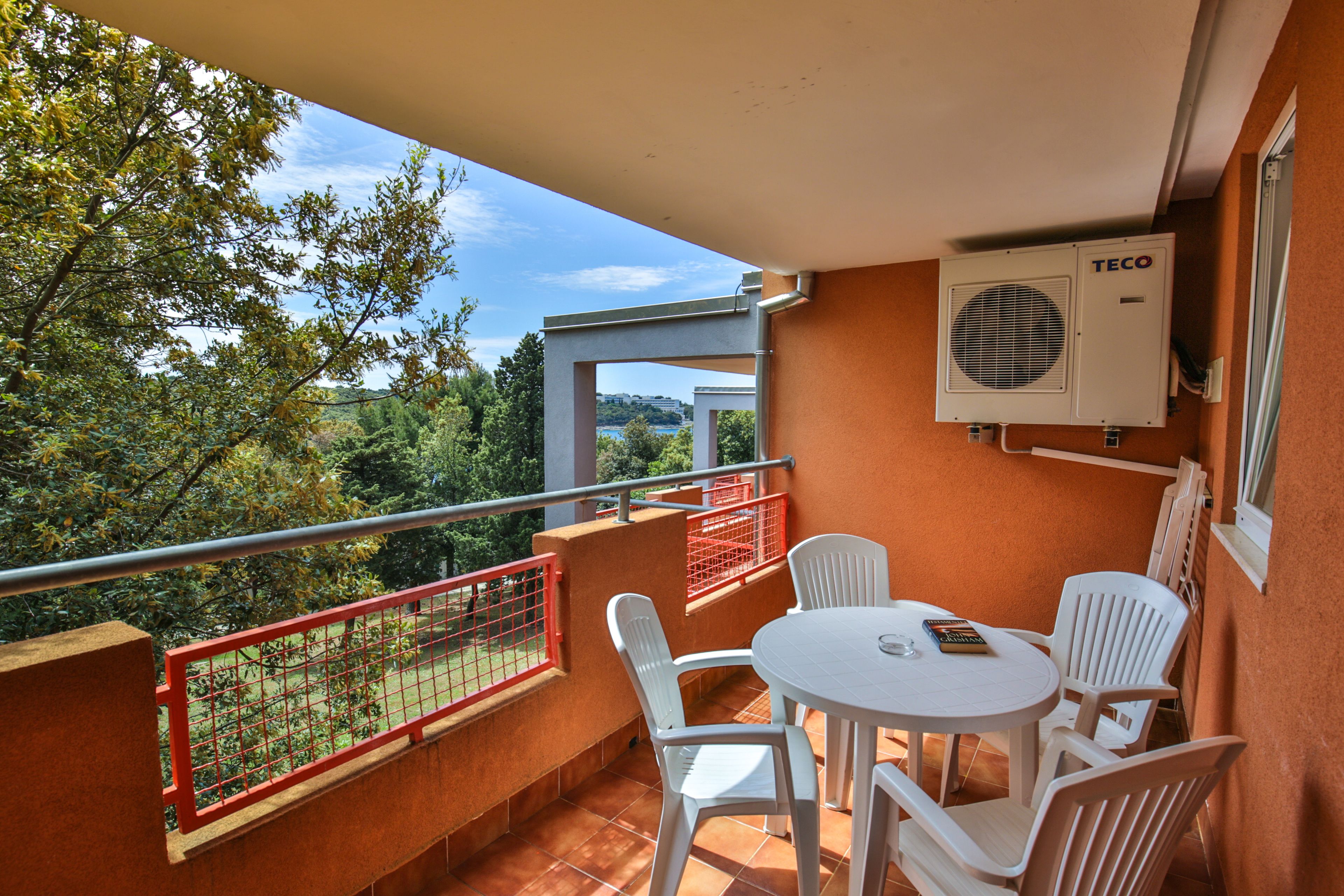 A balcony with a table and chairs, a red railing, and an air conditioning unit.