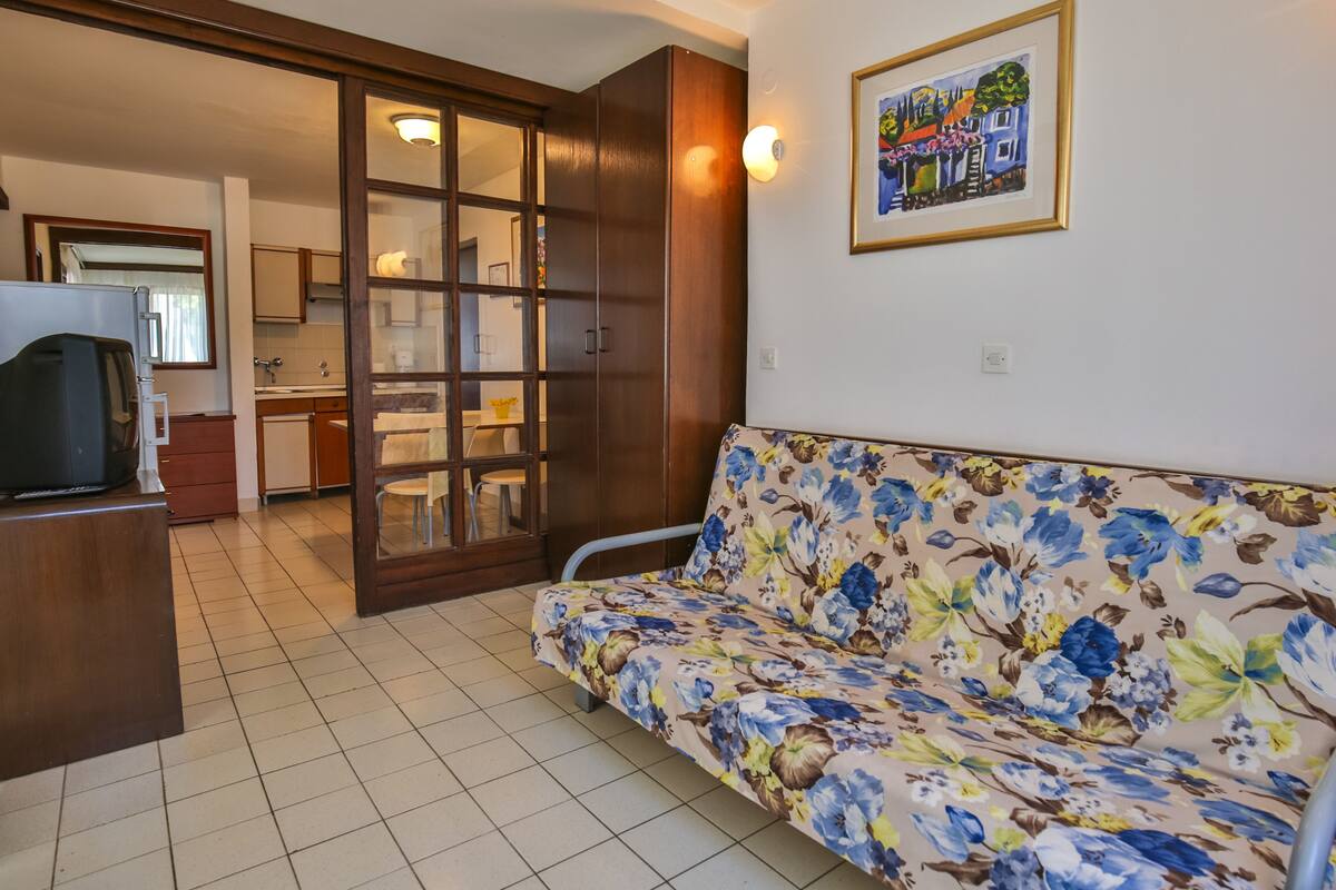 A living room with a floral sofa, a television, and a kitchen in the background.