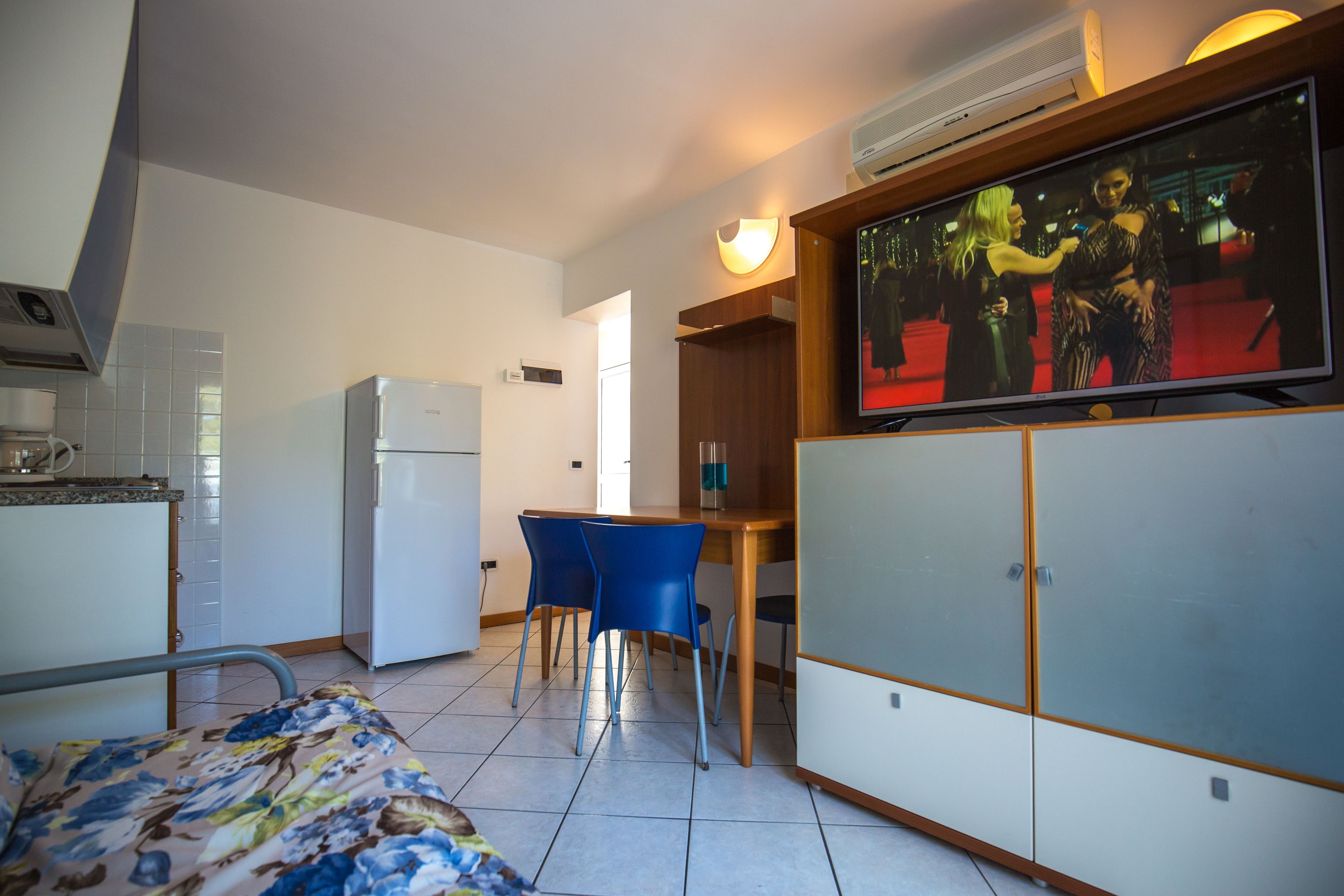 A compact hotel room with a bed, a kitchenette, a dining area with two chairs, and a TV displaying a red carpet event.