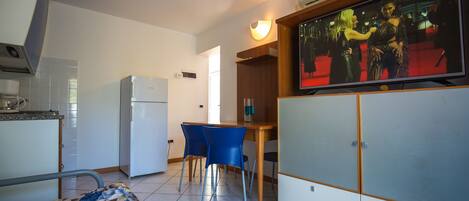 A compact hotel room with a bed, a kitchenette, a dining area with two chairs, and a TV displaying a red carpet event.
