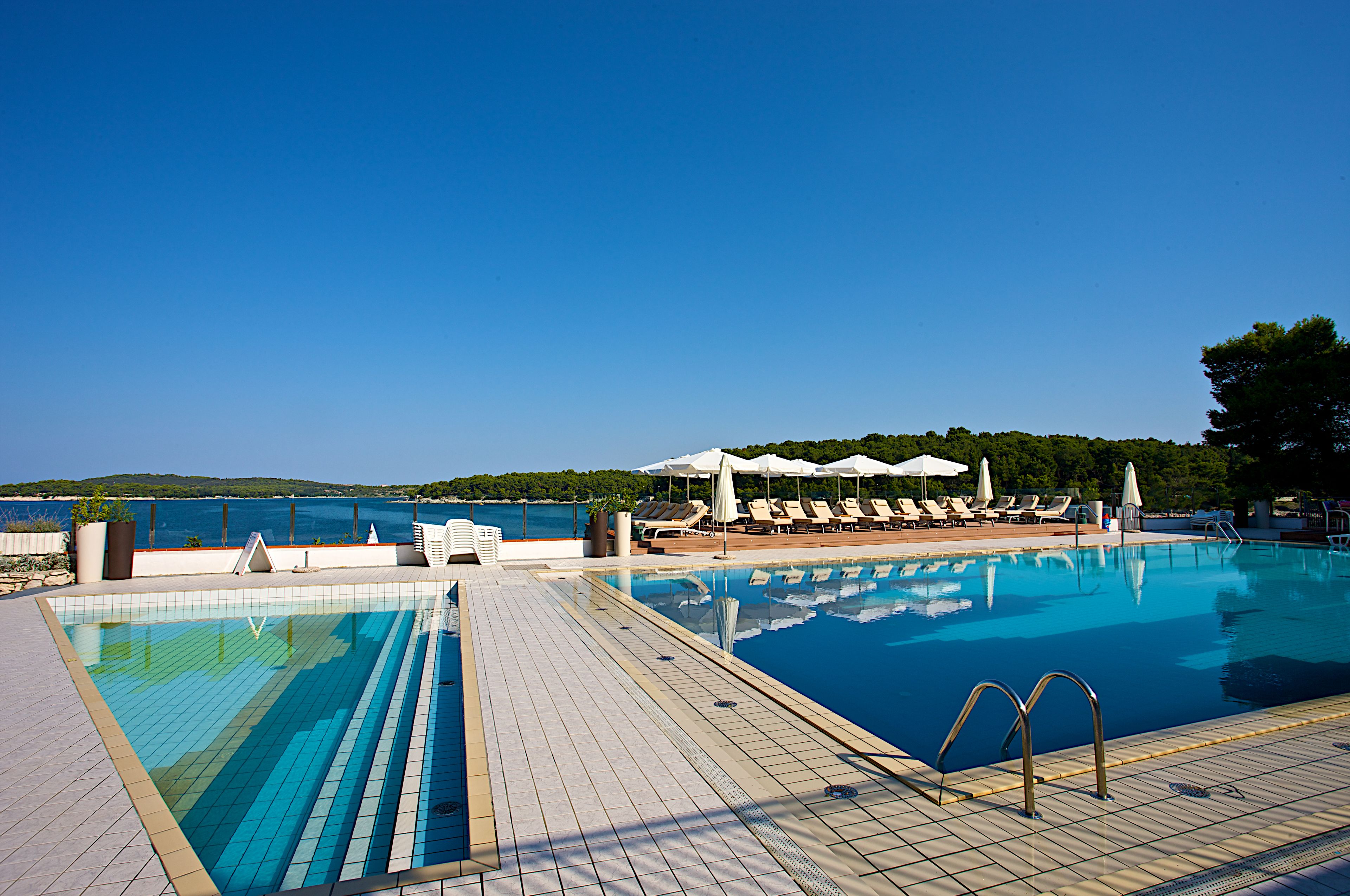 A swimming pool with lounge chairs and umbrellas, surrounded by a wooden deck and a view of the sea.