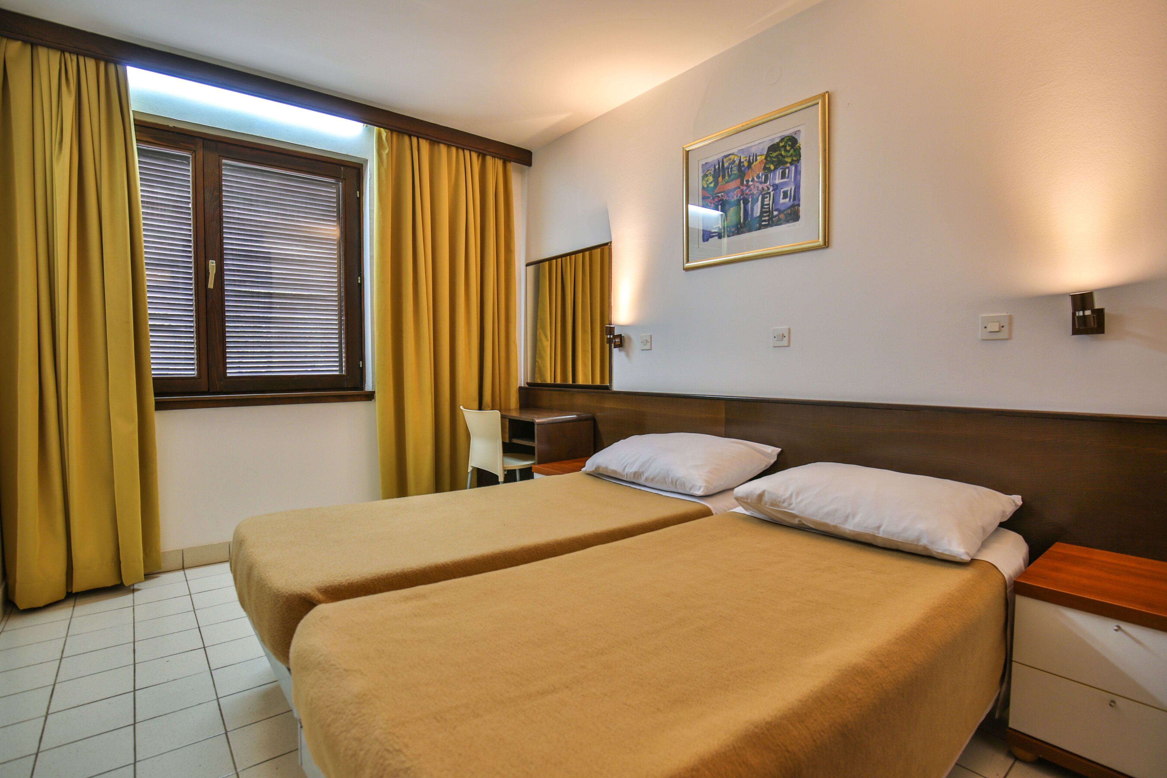 Une chambre d’hôtel avec deux lits, une fenêtre avec des stores, une chaise, une table de chevet et une lampe fixée au mur.