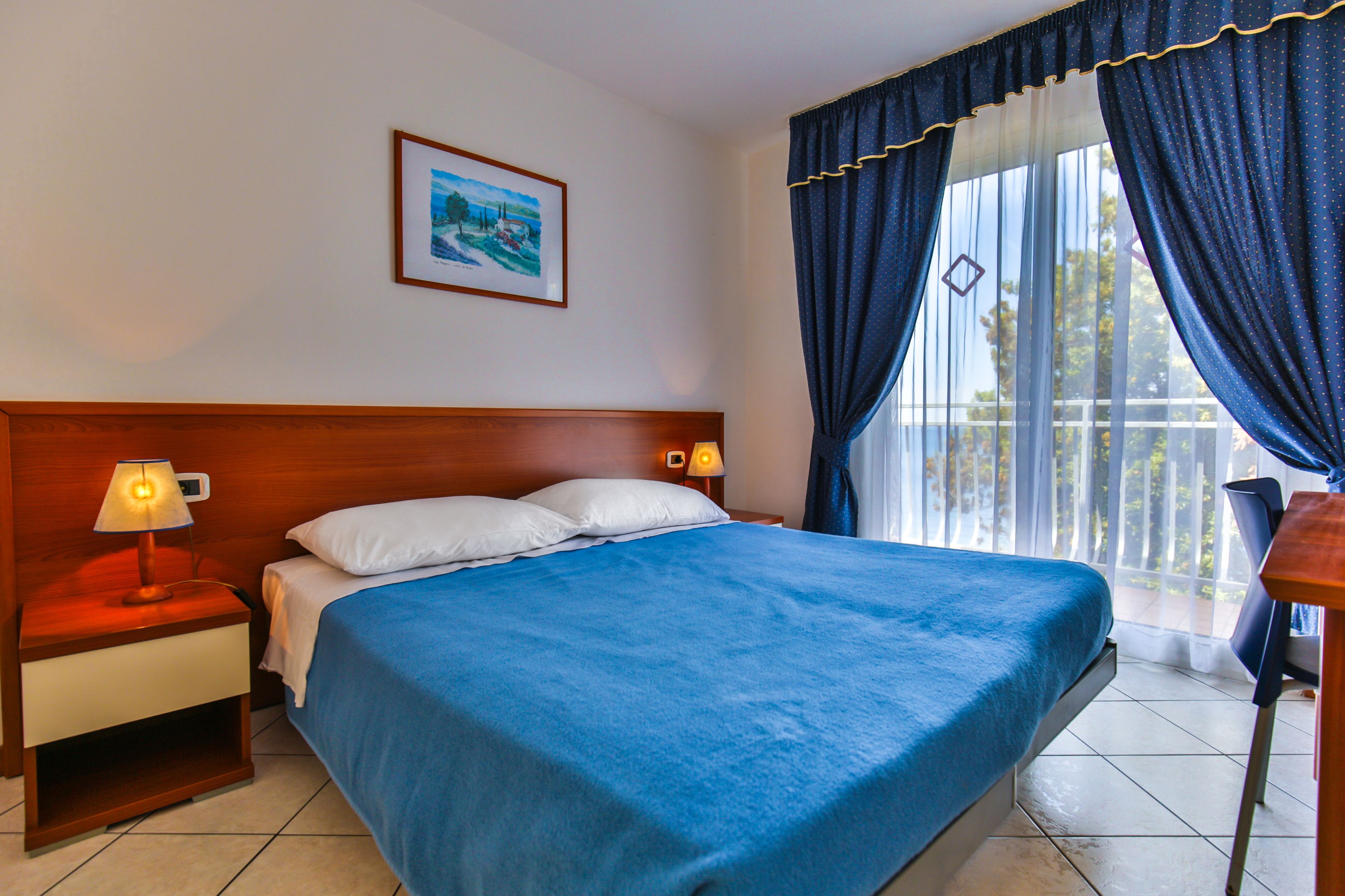 A hotel room with a bed, nightstand, lamp, and a framed picture on the wall.