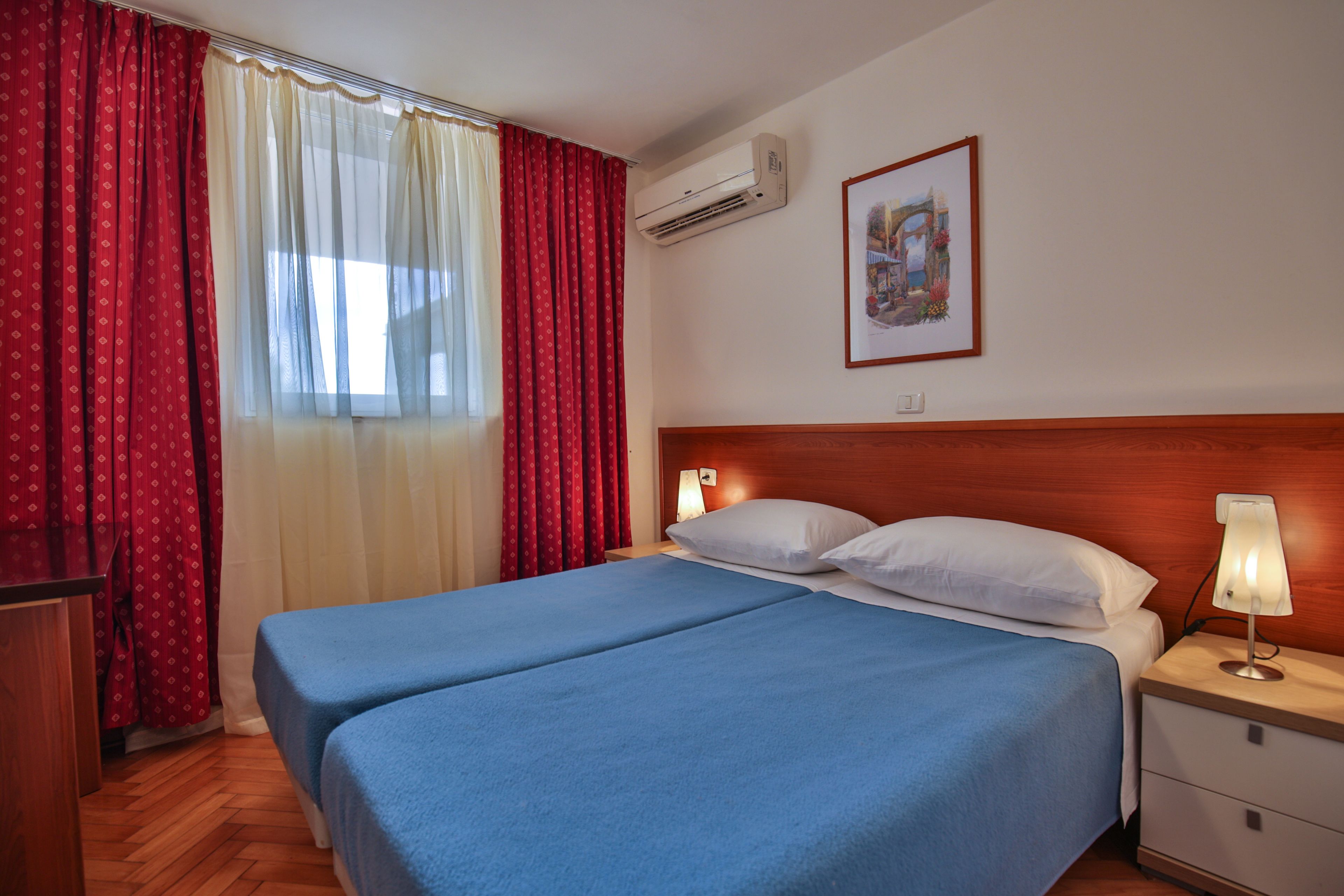 A hotel room with a double bed, a nightstand with a lamp, an air conditioner, and a framed picture on the wall.
