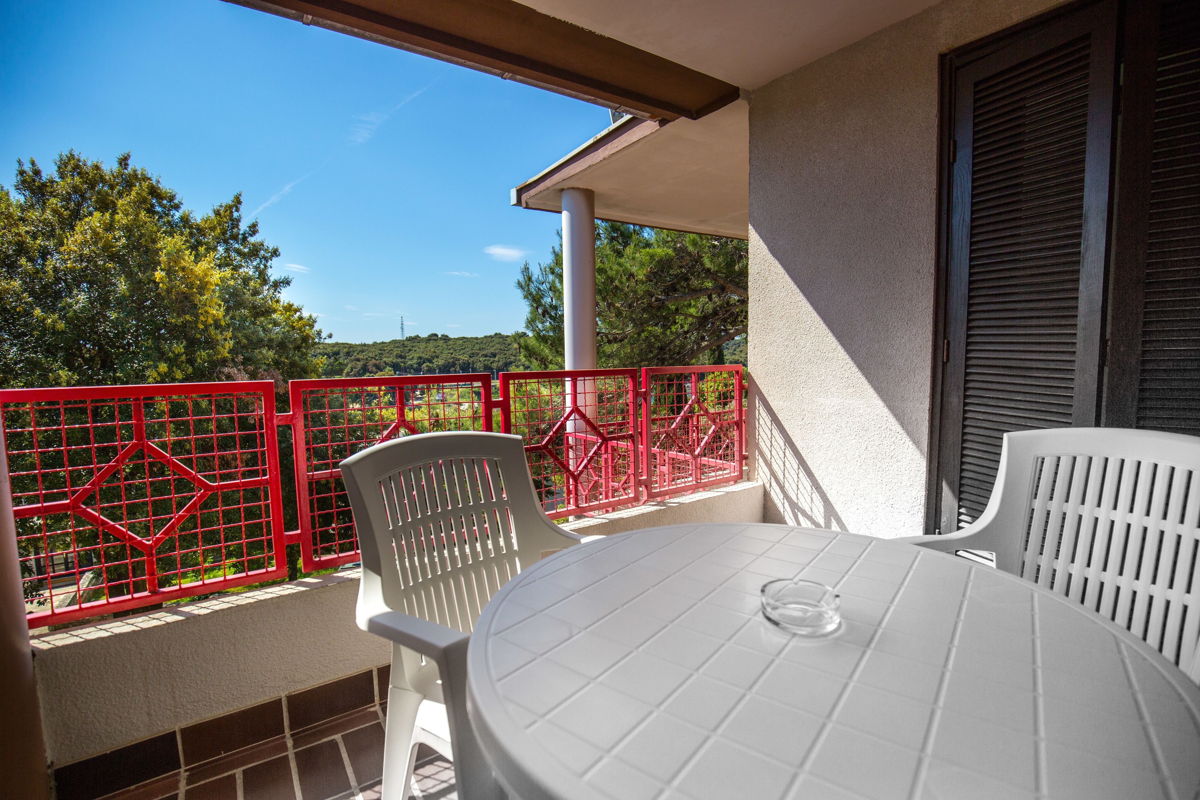 A balcony with a white table and chairs, a red metal fence, and a view of trees and hills.