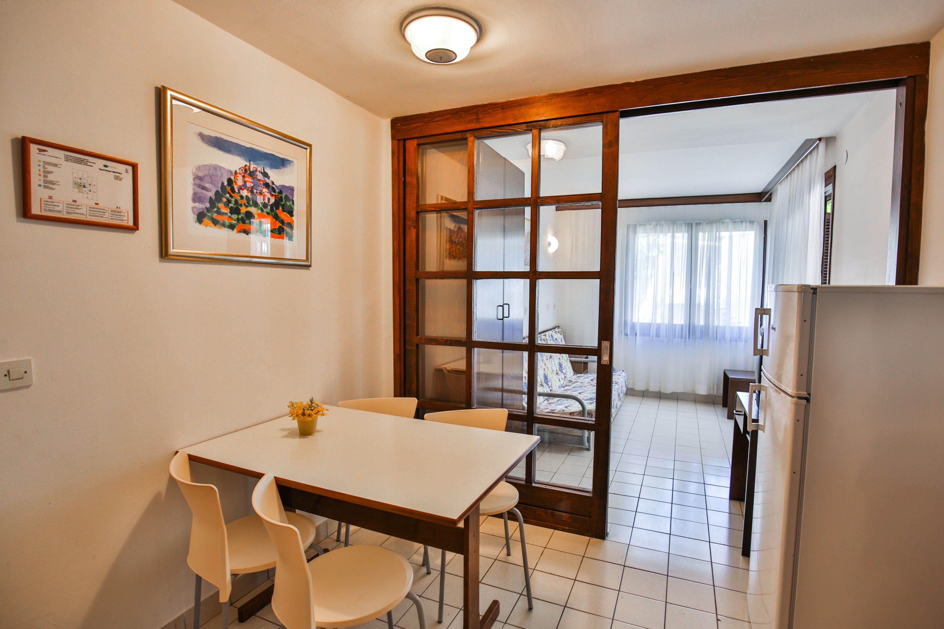 A small kitchen with a dining table and chairs, a refrigerator, and a sliding glass door leading to a living area.