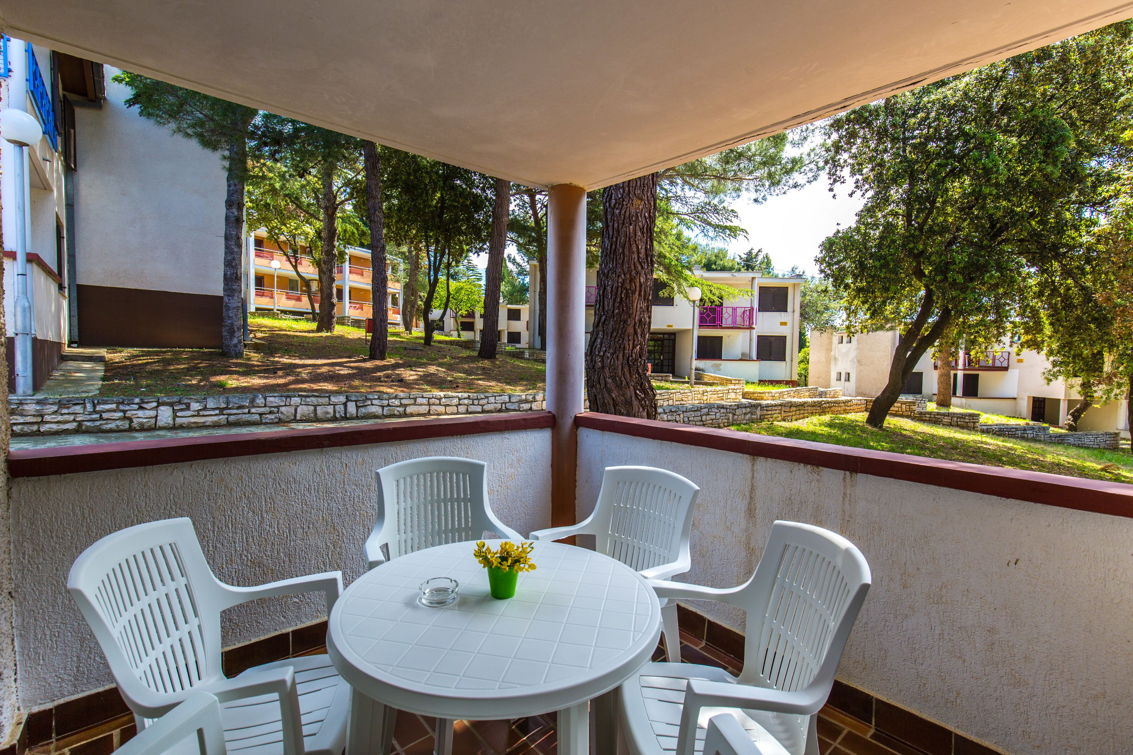 Zona salotto all'aperto coperta, con tavolo e sedie, affacciata su un cortile con alberi e edifici.
