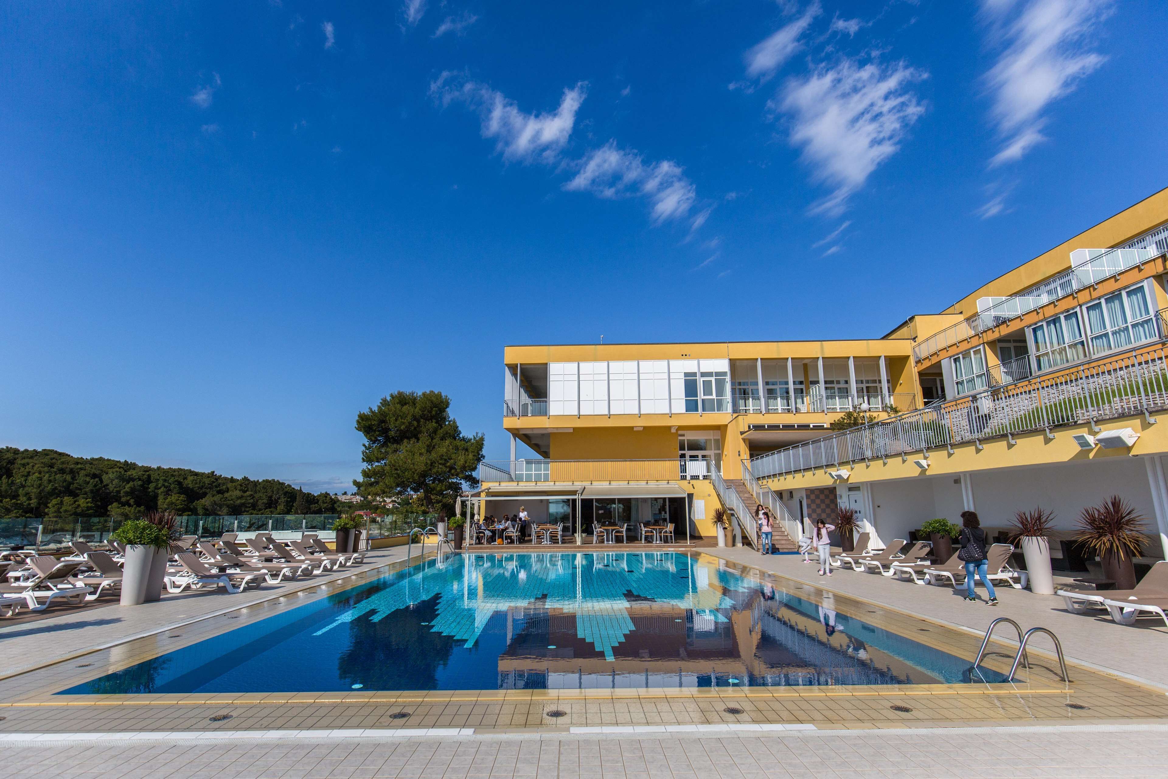 Ein Poolbereich mit Liegestühlen, ein Gebäude mit Balkonen und ein klarer, blauer Himmel.