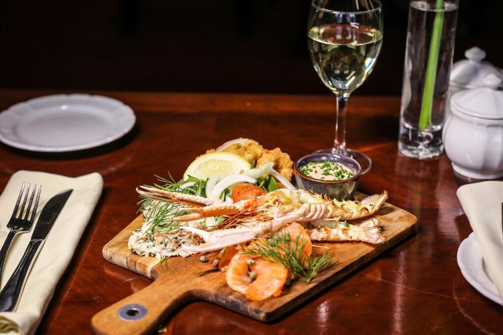 Un plato de mariscos sobre una tabla de madera, con una copa de vino blanco, un recipiente pequeño de salsa y un salero en una mesa de madera.