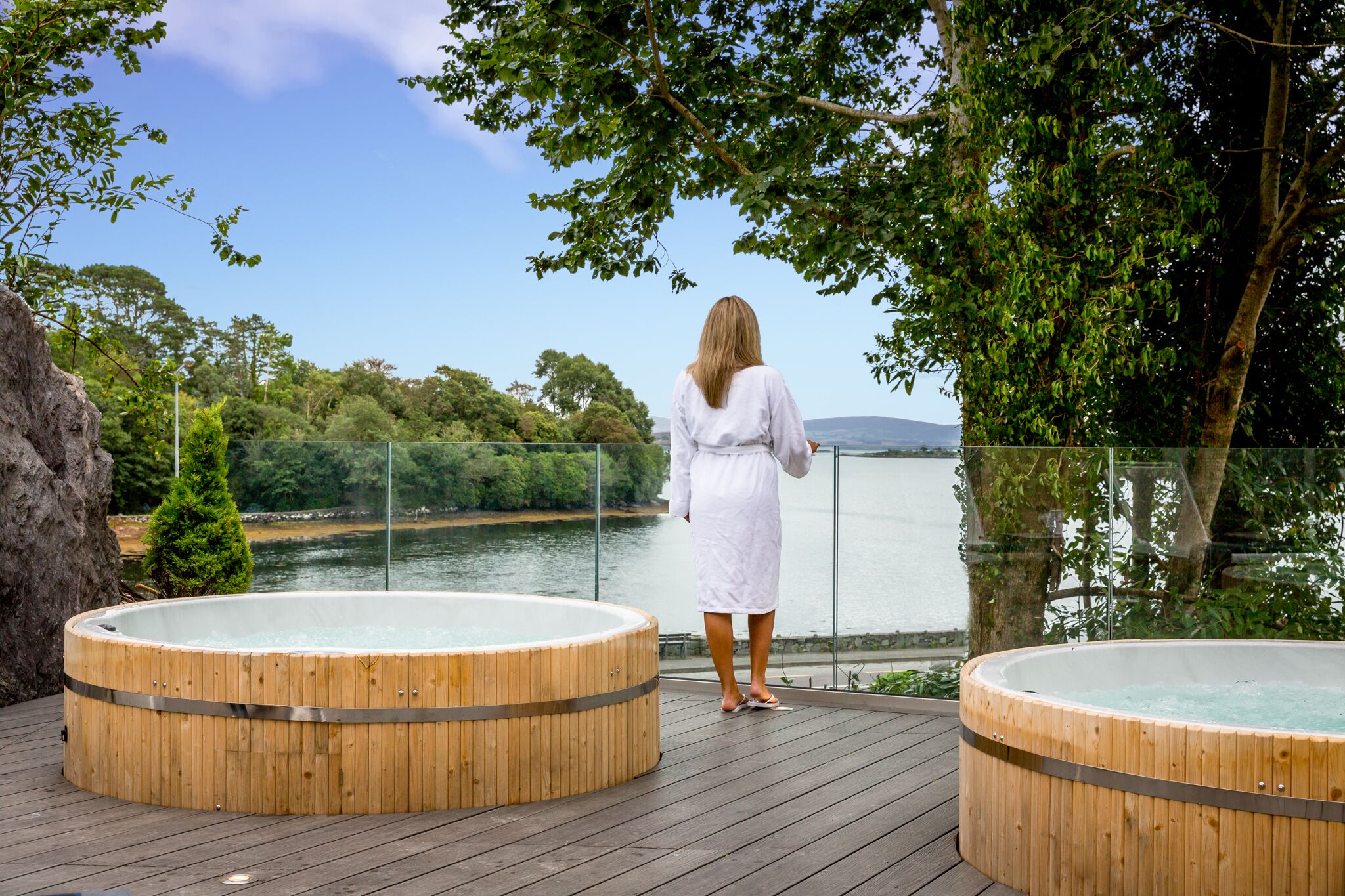 A woman in a white dress standing between two wooden hot tubs on a wooden deck overlooking a lake with trees and hills in the background.