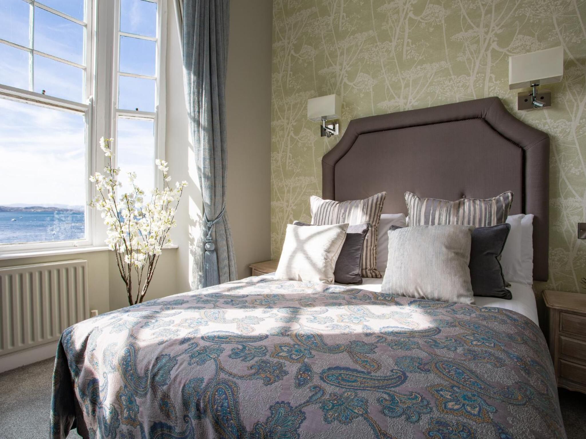 A bedroom with a large bed, a view of the sea through a window, and a vase with flowers.