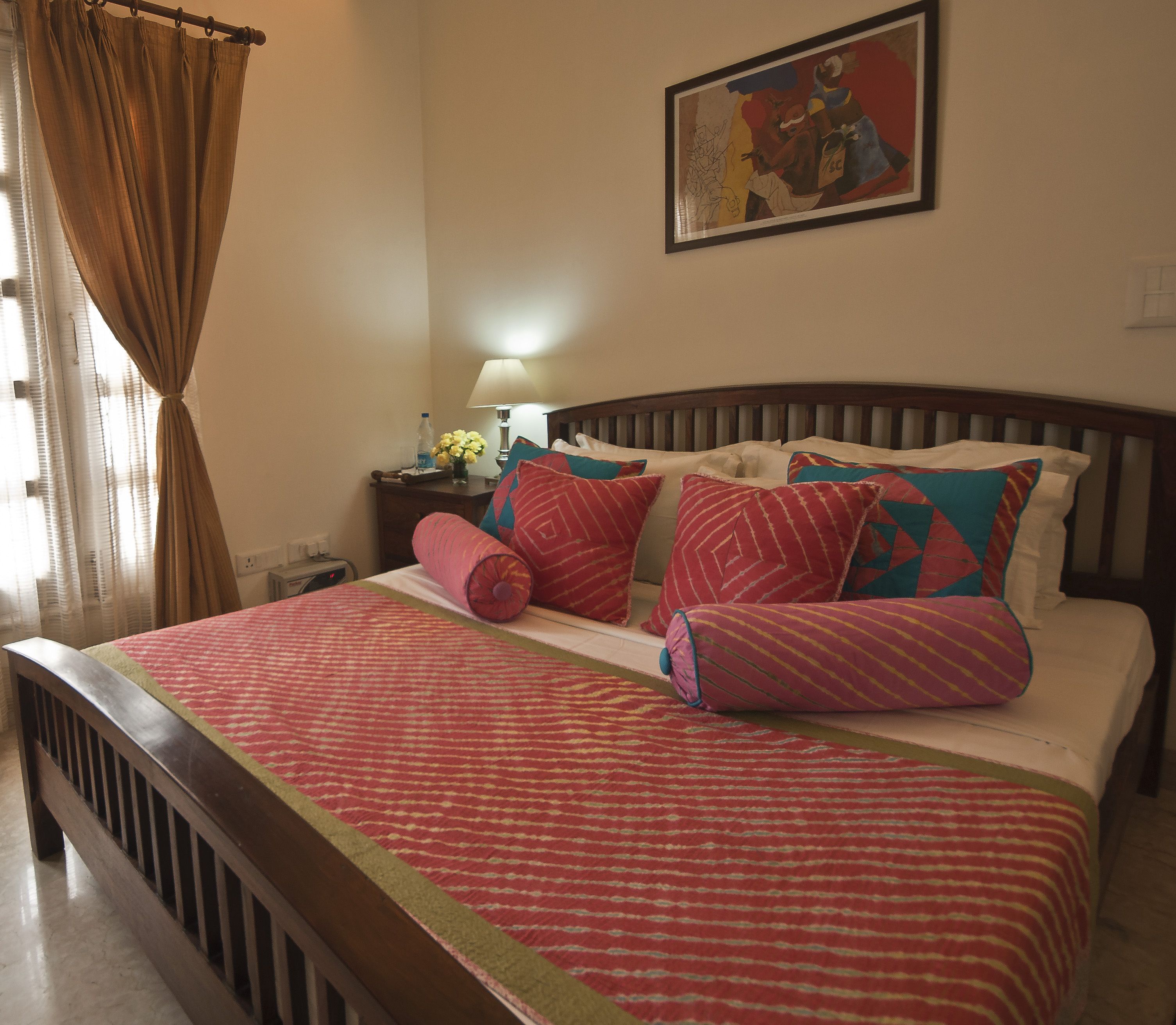 A bed with a patterned bedspread, multiple pillows, a nightstand with a lamp, and a painting on the wall.