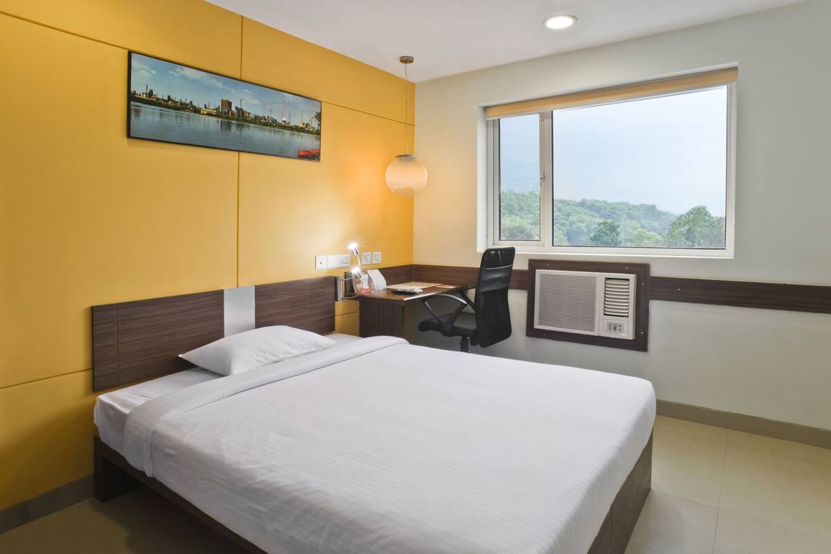 Habitación de hotel con una cama grande, un escritorio con una silla, una ventana con vista a follaje y un cuadro en la pared.