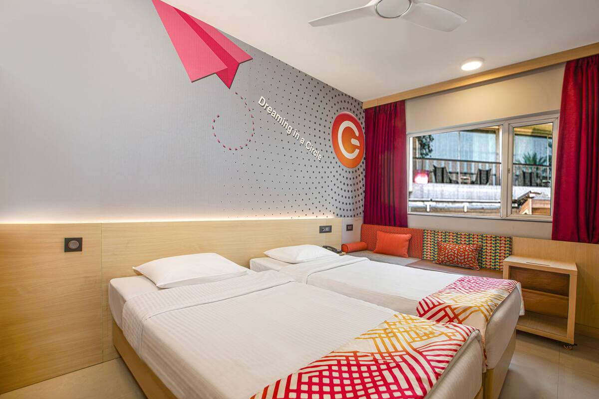 Habitación de hotel con dos camas, un escritorio, un ventanal con cortinas rojas y una pared con un diseño circular y texto.