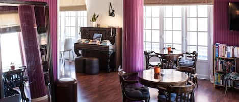 A room with a piano, a dining table with chairs, a bookshelf, and large windows.