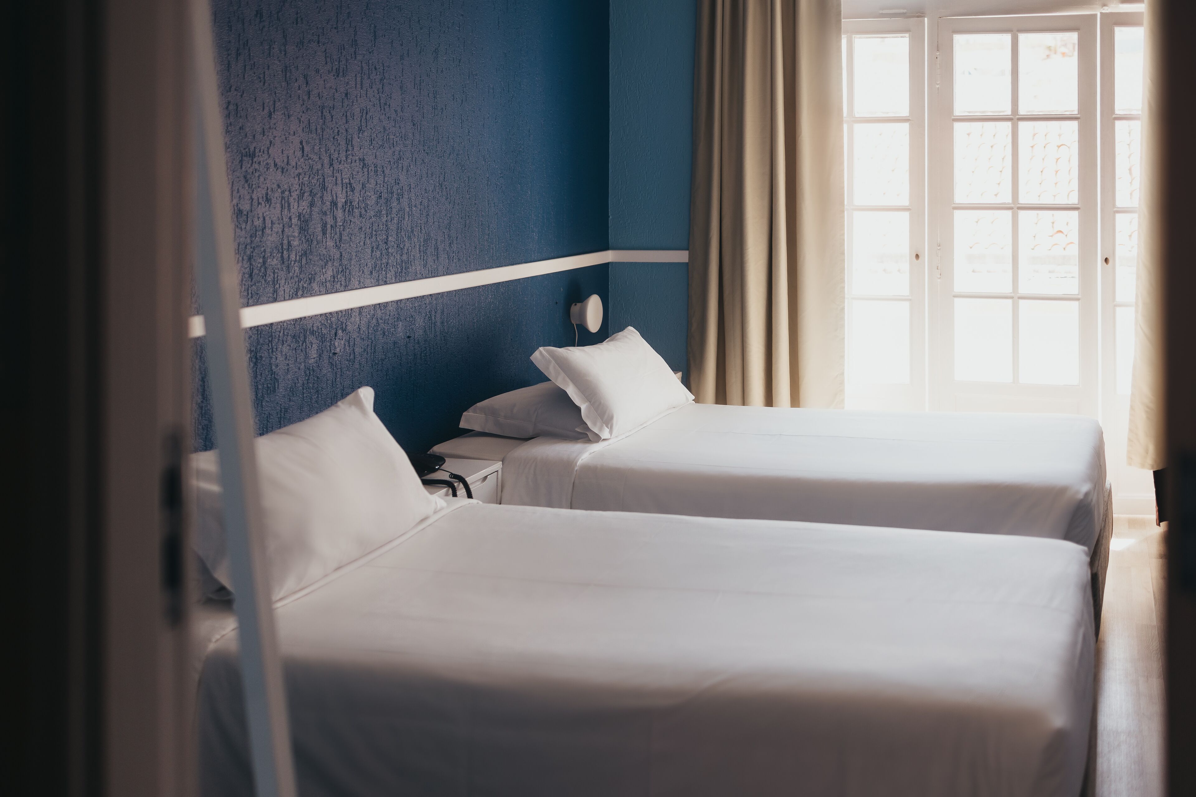Habitación de hotel con dos camas, una pared azul decorativa y una ventana con cortinas.