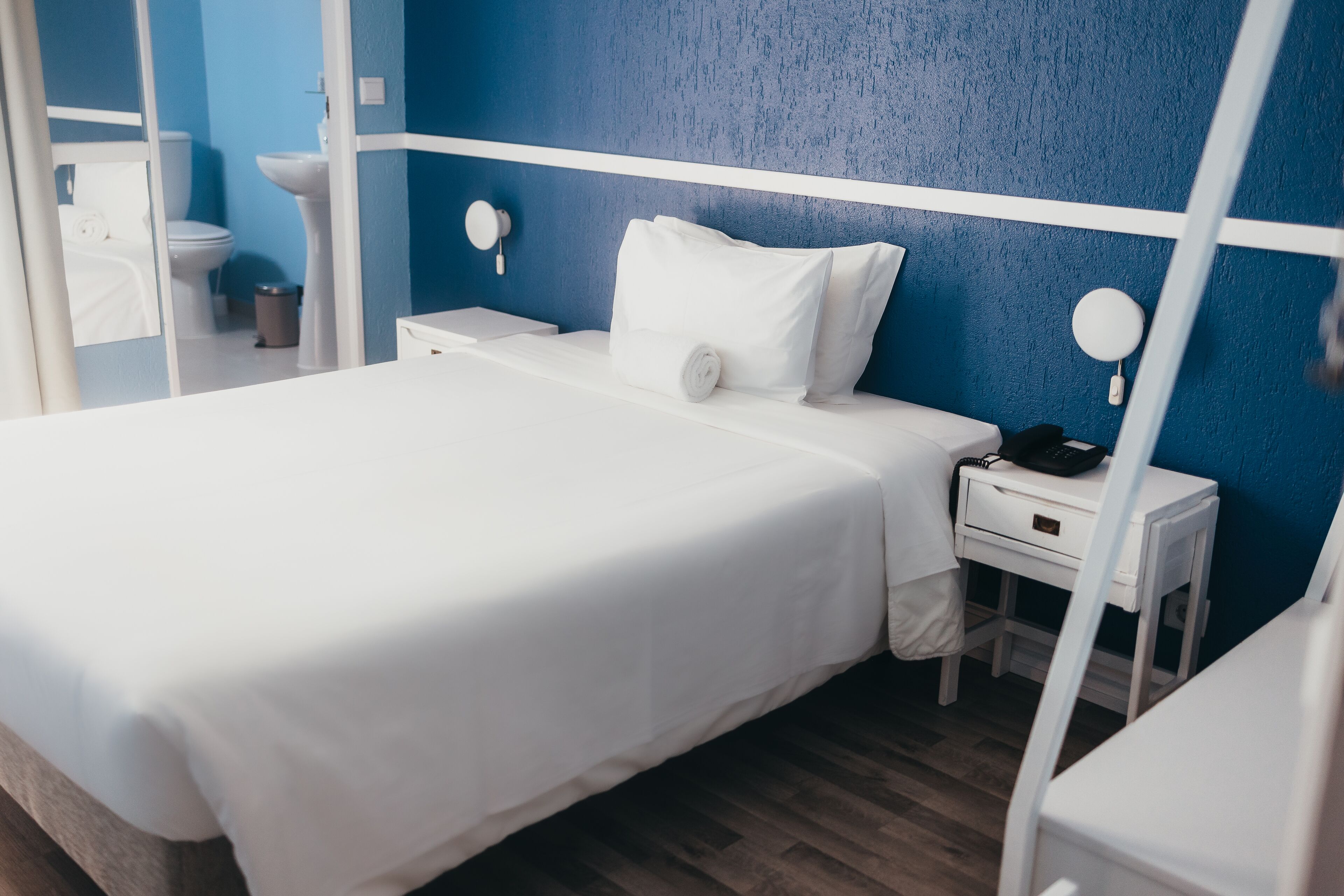 A neatly made bed with white bedding, a bedside table with a lamp, and a blue textured wall.