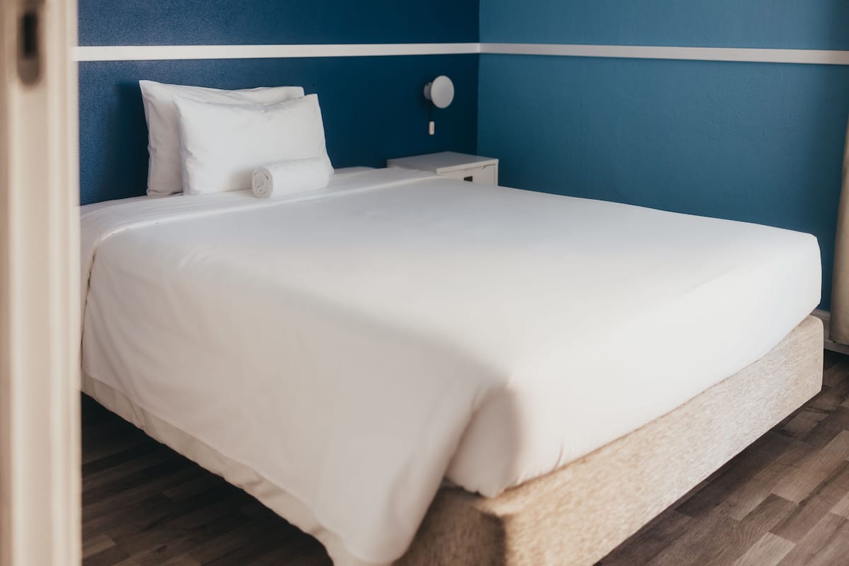 Ein ordentlich bezogenes Bett in einem Zimmer mit blauer Akzentwand, einem kleinen Nachttisch und Holzboden.