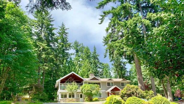 A two-story house with a stone foundation, surrounded by lush greenery and tall trees. - Maple View Bed and Breakfast (Nanaimo)