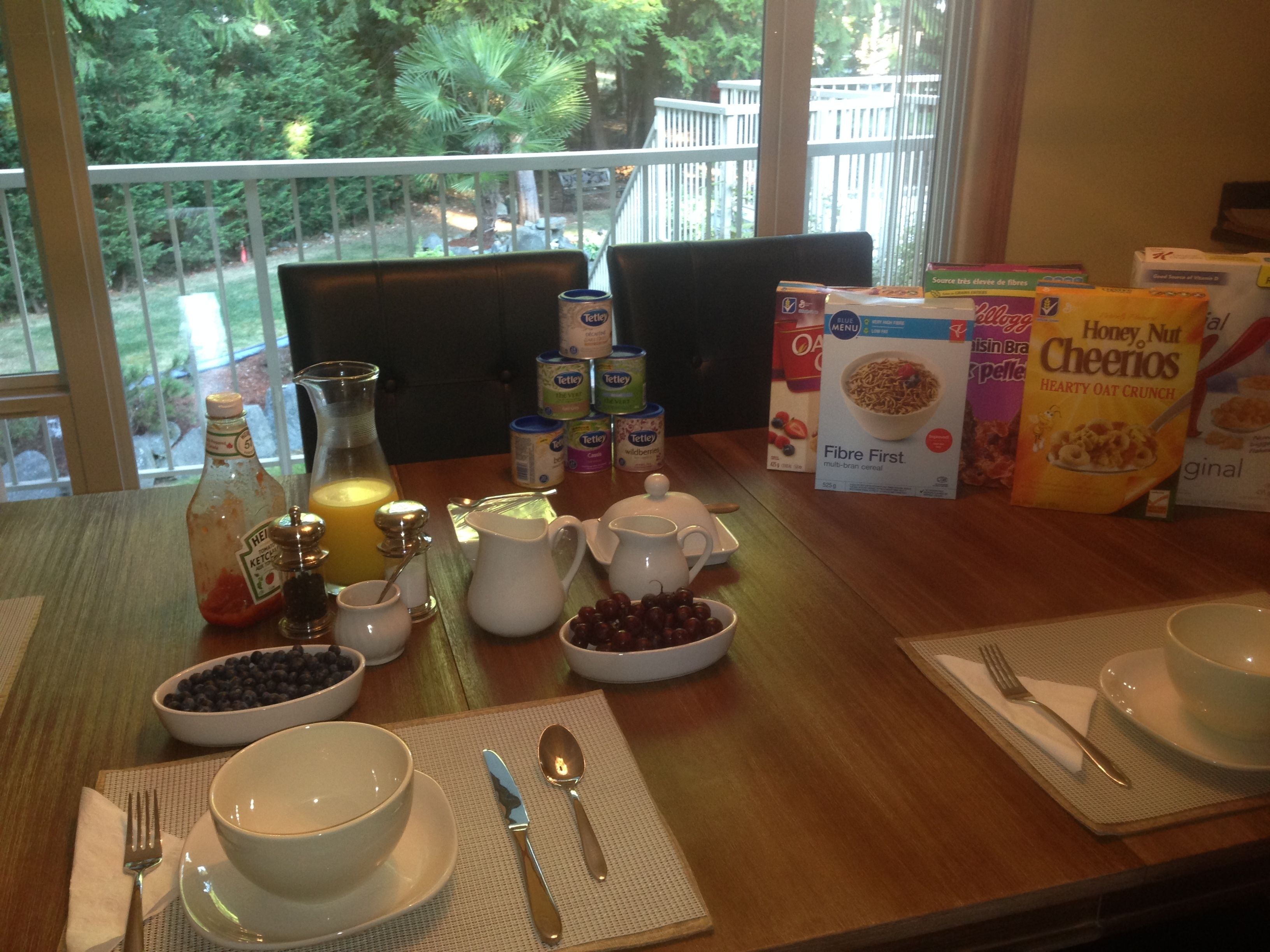 A breakfast table with various food items, including cereal boxes, a bottle of ketchup, and bowls of fruit.