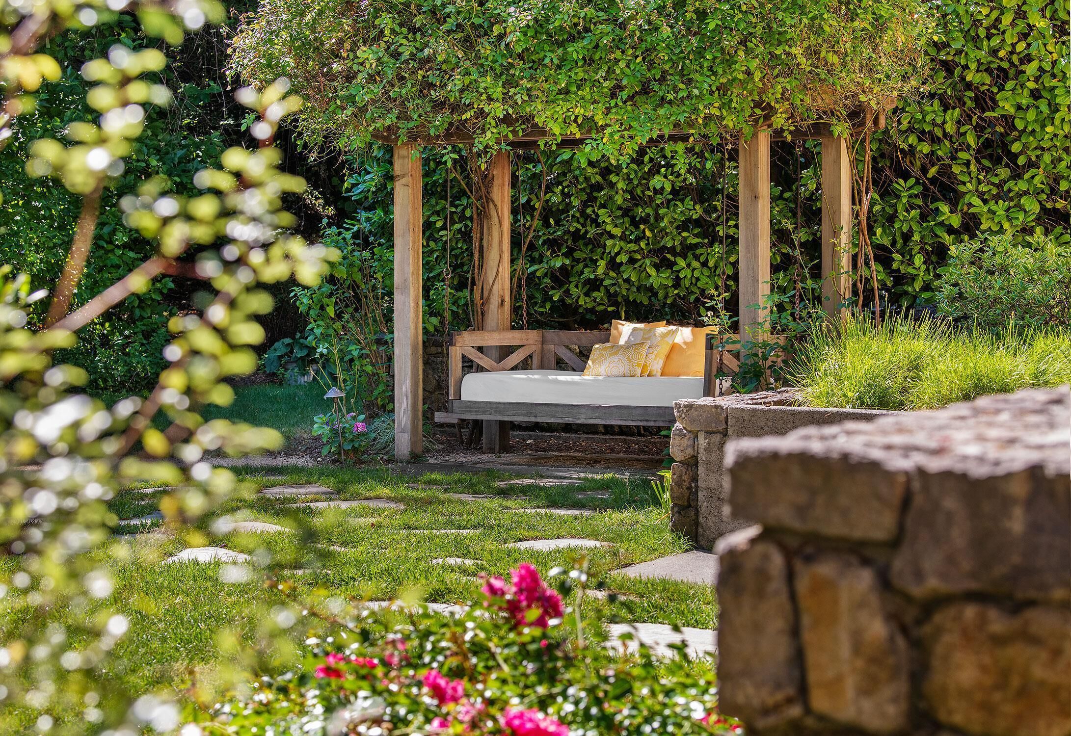 A garden with a wooden swing bed, surrounded by greenery and stone structures.