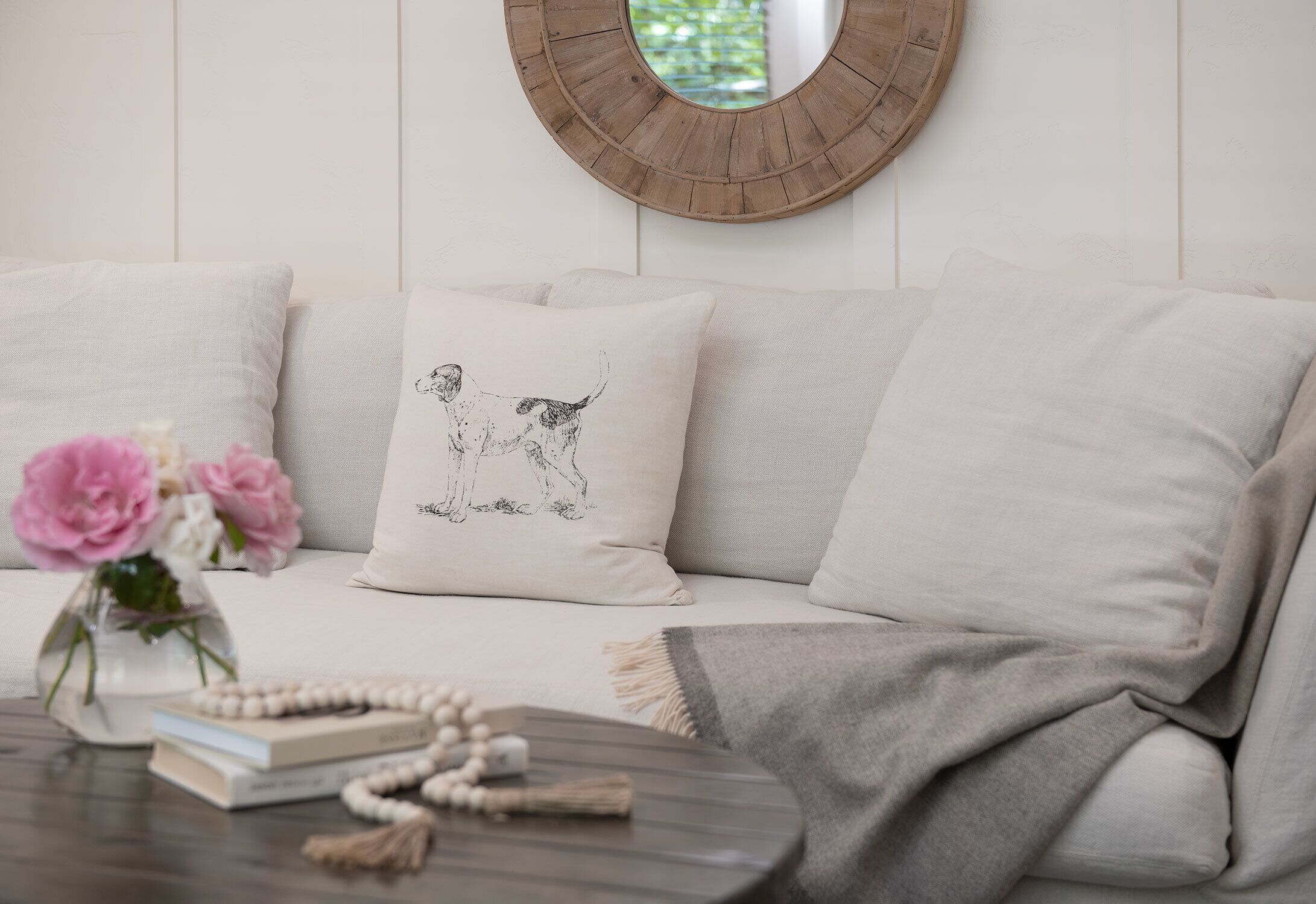 A neutral-toned sofa with a single decorative pillow featuring a dog illustration, a round mirror on the wall, and a small table with books and a rosary.