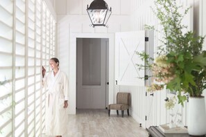A person in a robe standing near a white door with a view of a room with a chair and a plant. - Farmhouse Inn (Forestville)
