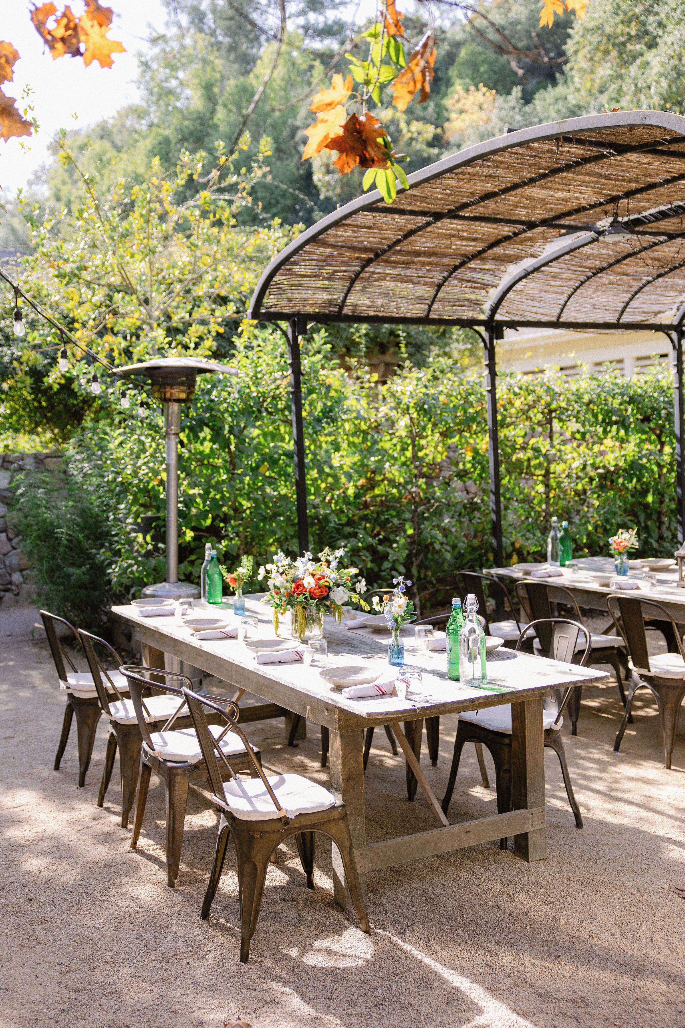 An outdoor dining area with tables and chairs, surrounded by greenery and a pergola structure.