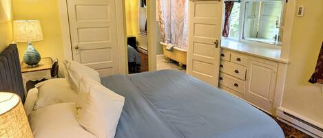 A bedroom with a bed, a nightstand with a lamp, a dresser, and a window with curtains.