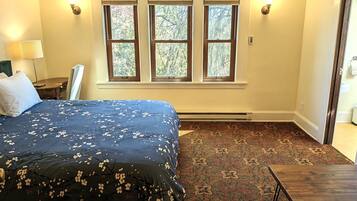 A bedroom with a large bed, a small table, and three windows with blinds.