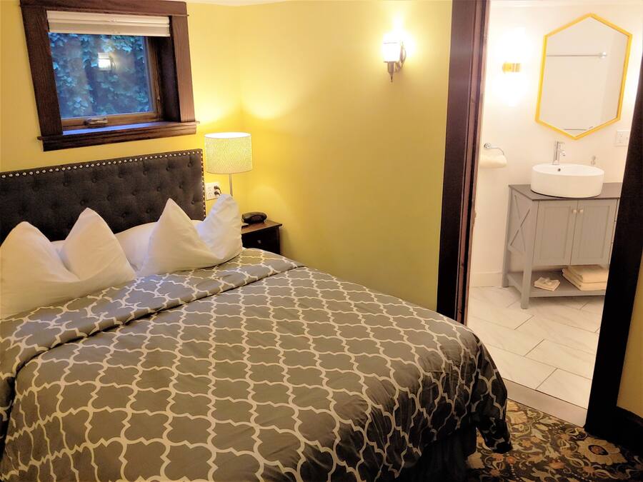 Une chambre à coucher avec une tête de lit capitonnée, un lit avec une literie à motifs, une table de chevet avec une lampe, et une vue sur une salle de bain équipée d’un lavabo et d’un miroir.