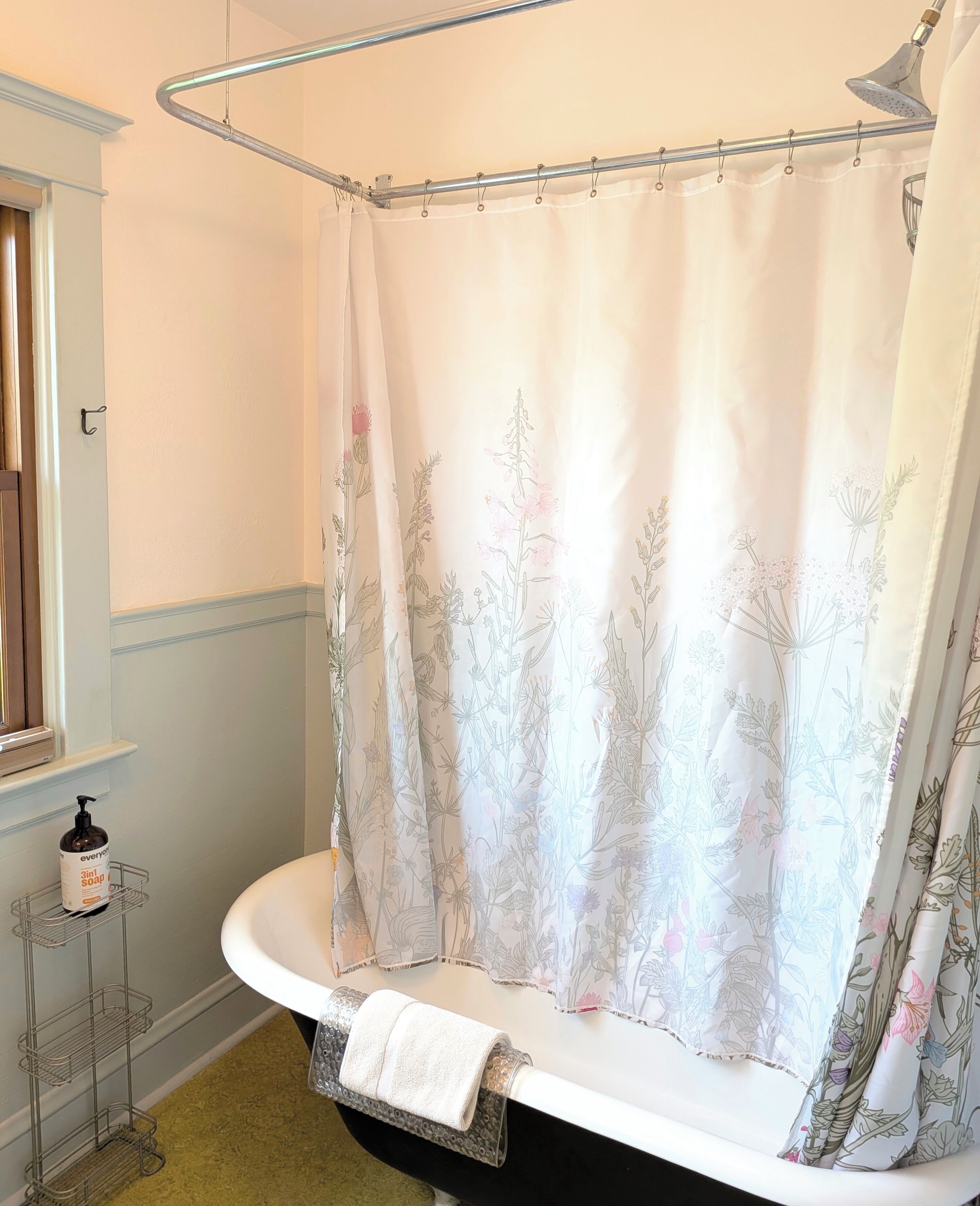 Un baño con cortina de ducha estampada con motivos florales, un lavabo blanco y un dispensador de jabón sobre una repisa metálica.