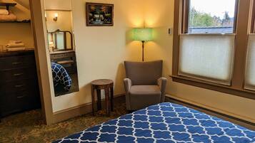 Une chambre d’hôtel avec un lit, une chaise, une petite table, une lampe, un tableau et une fenêtre avec des stores.