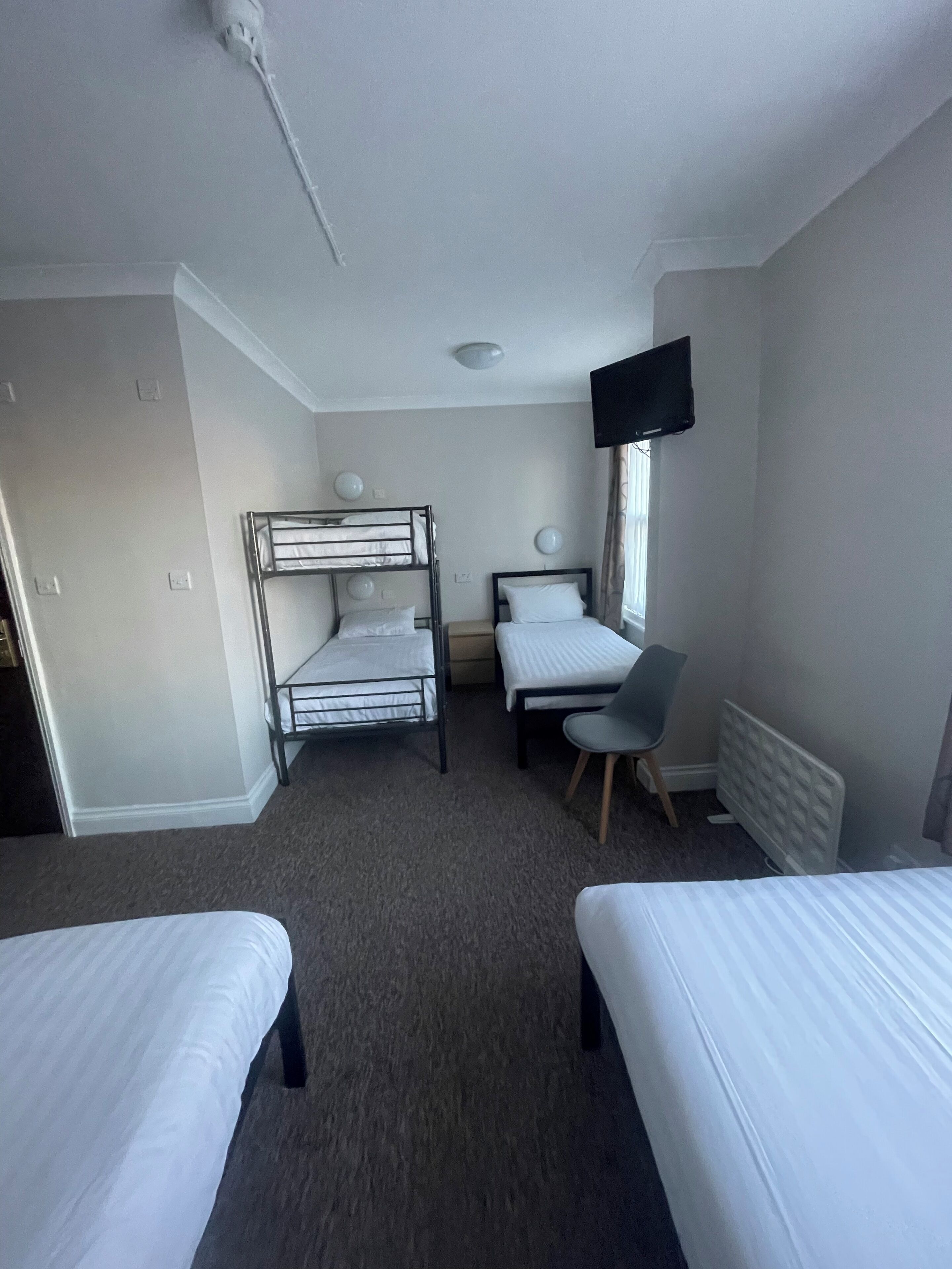 A hotel room with two single beds, a bunk bed, a desk with a chair, a flat-screen TV, and a radiator.