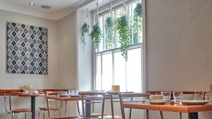 Un comedor con mesas de madera y sillas de metal, ventanas amplias con plantas colgantes y un muro con diseño.