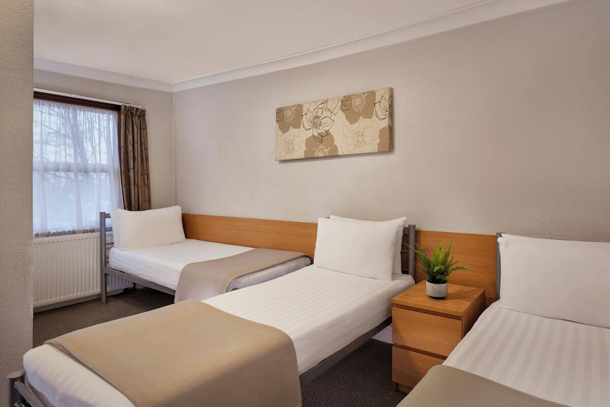 Habitación de hotel con tres camas individuales, cabecera de madera, una planta en maceta y una obra de arte floral grande en la pared.