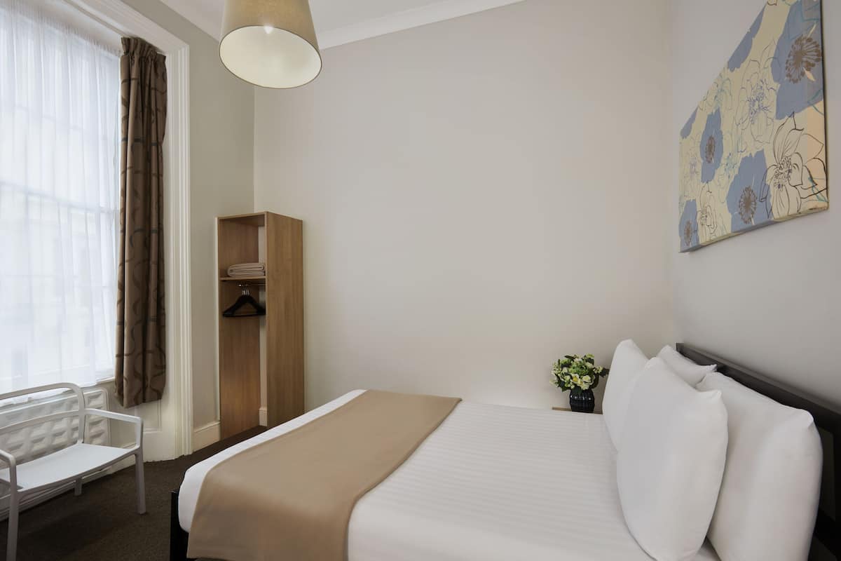 A hotel room with a bed, a chair, a wardrobe, and a painting on the wall.