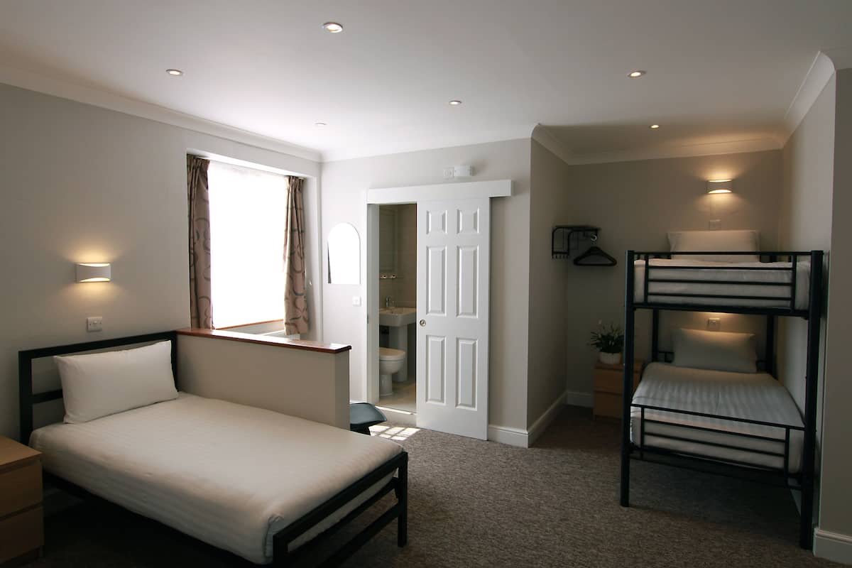 Un dormitorio con una cama individual, camas literas, una mesita de noche, un escritorio, una silla, una ventana con cortinas, una puerta y una lámpara fijada a la pared.
