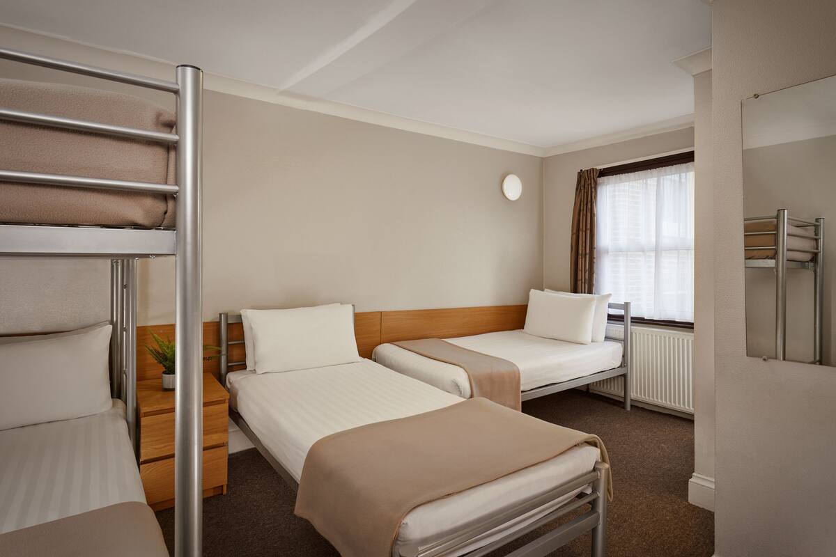 Habitación de hotel con dos camas individuales, una litera, una mesita de noche con una planta, una ventana con cortinas y un radiador.