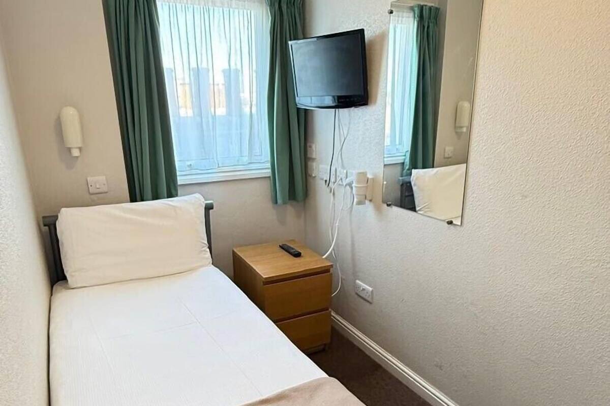 A hotel room with a bed, a bedside table, a TV, a mirror, and a window with curtains.