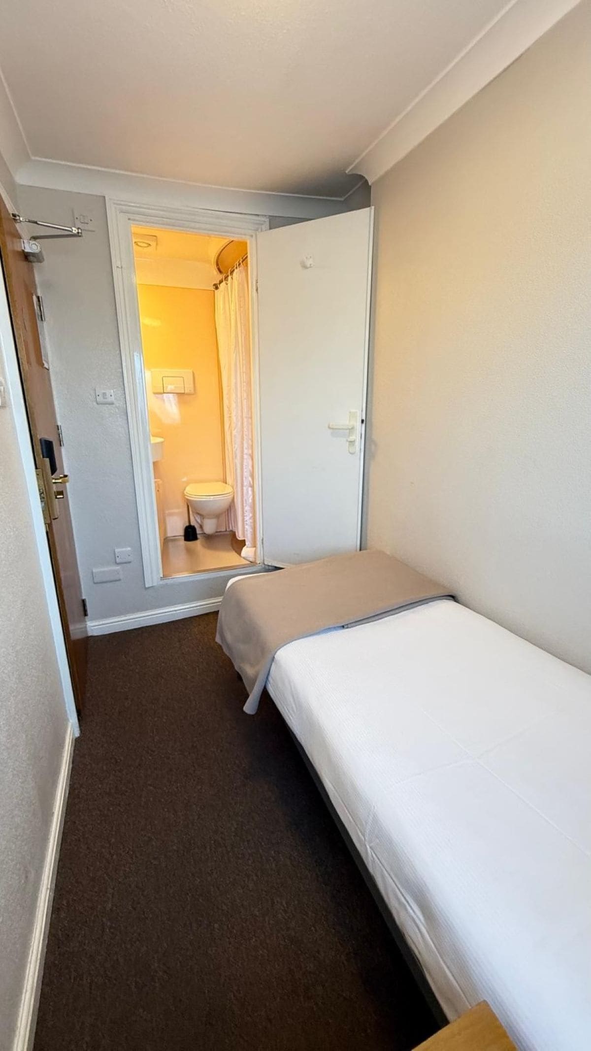 Habitación de hotel estrecha con una cama individual, baño pequeño con inodoro y ducha, y una puerta que da a otra habitación.