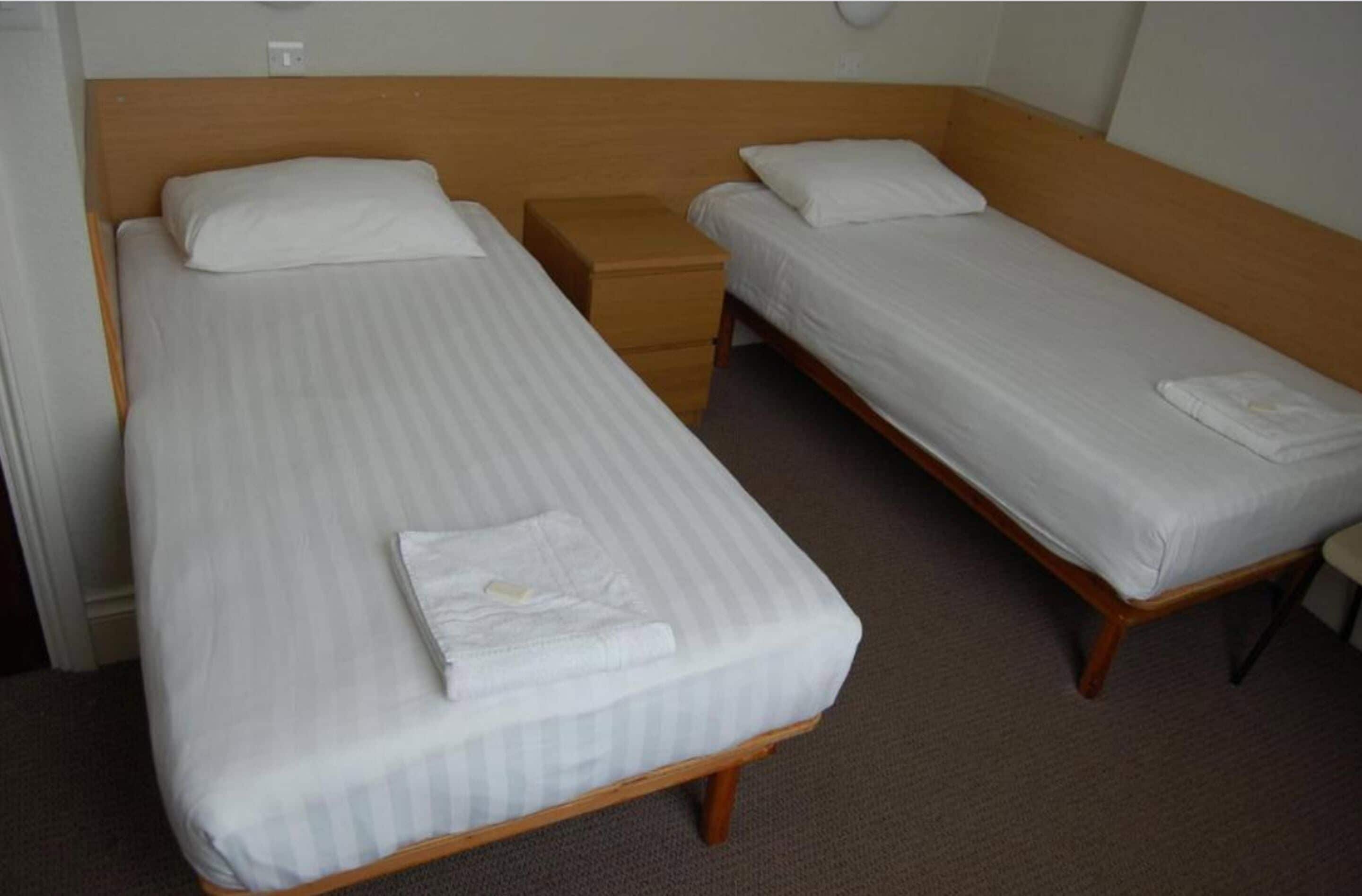 A room with two single beds, each with a wooden headboard and white bedding. There is a small wooden nightstand between the beds.