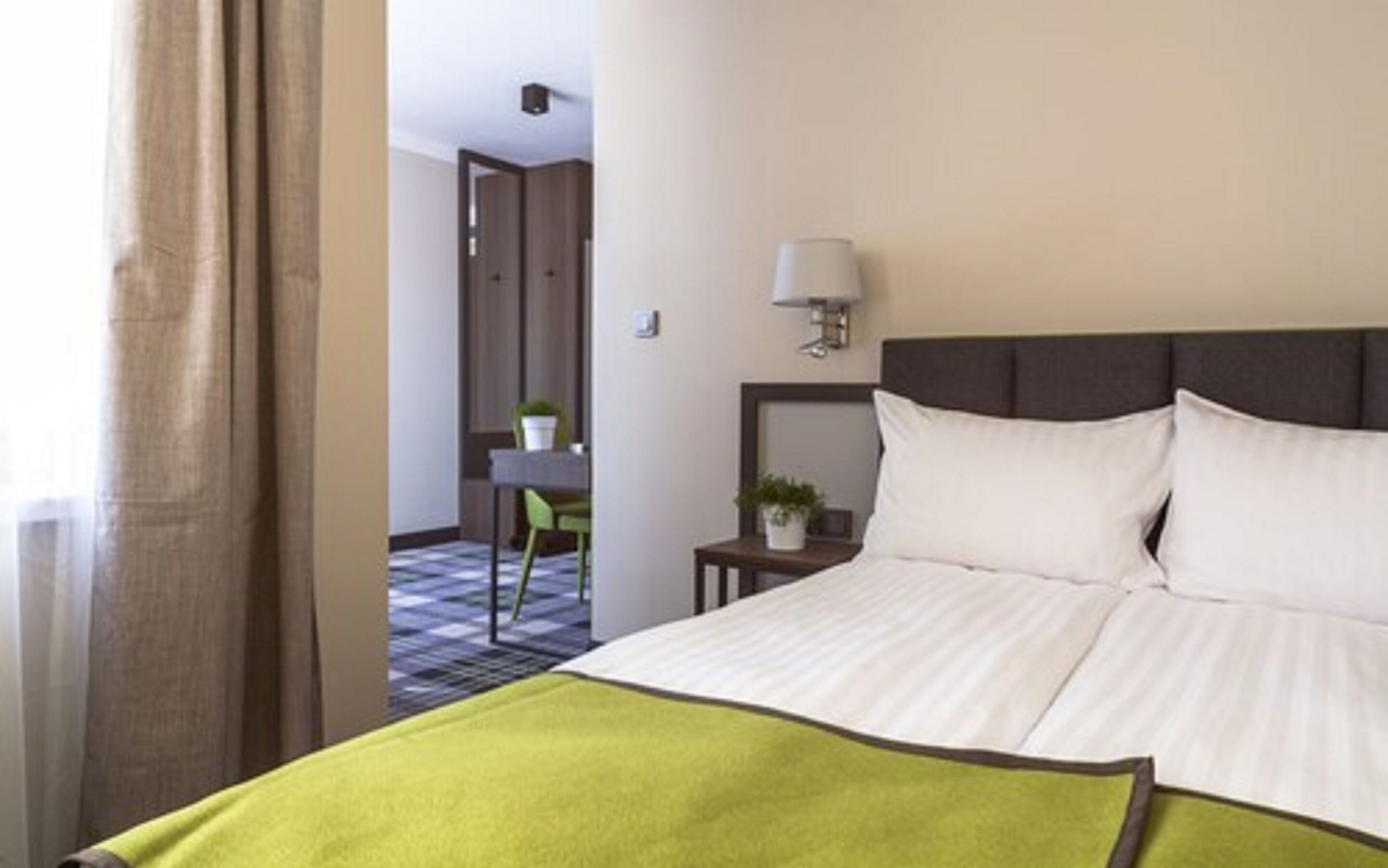A hotel room with a bed, a nightstand, a lamp, and a view of a hallway with a door and a chair.
