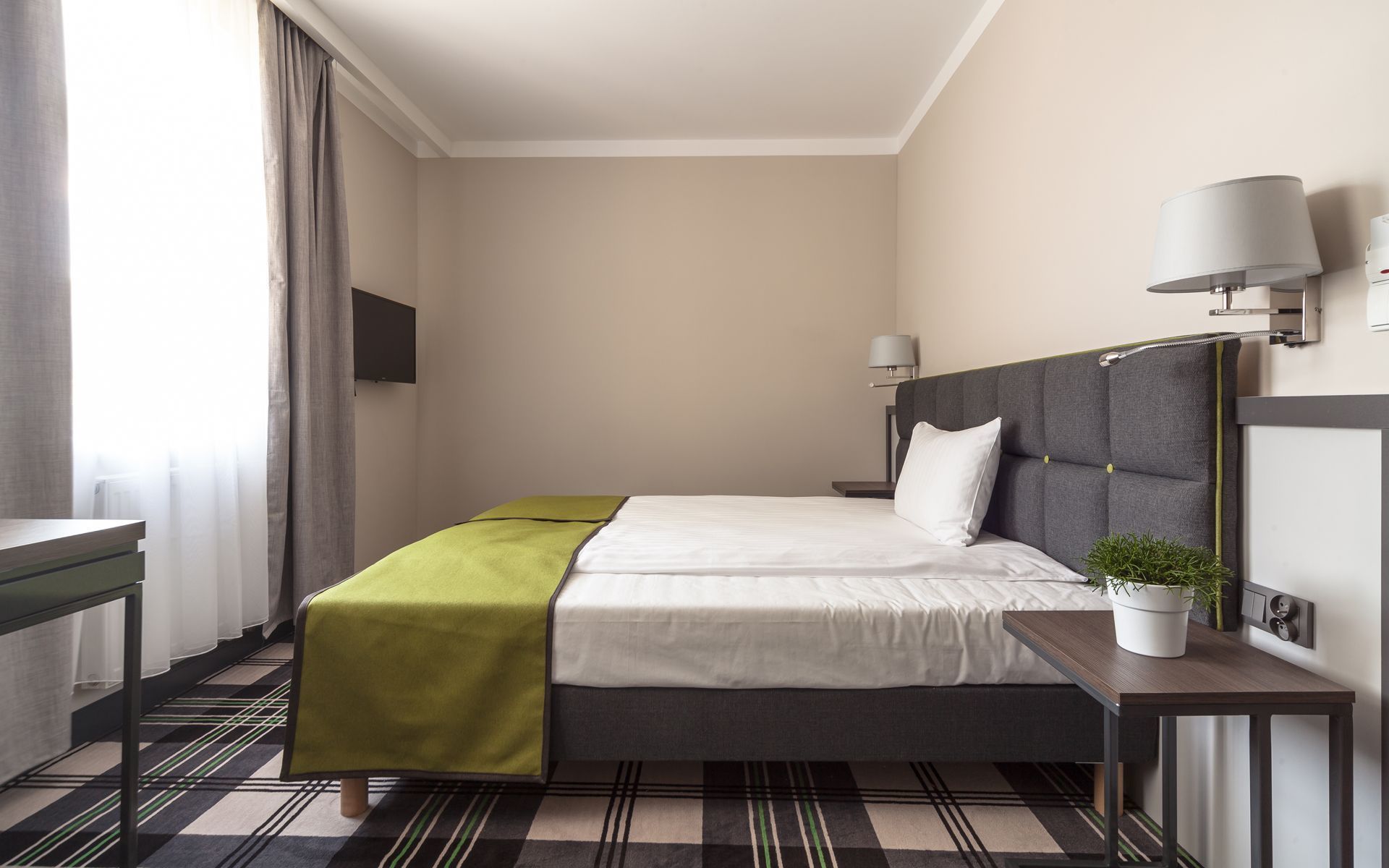 Ett modernt hotellrum med en säng, sängbord, en tv och en krukväxt.